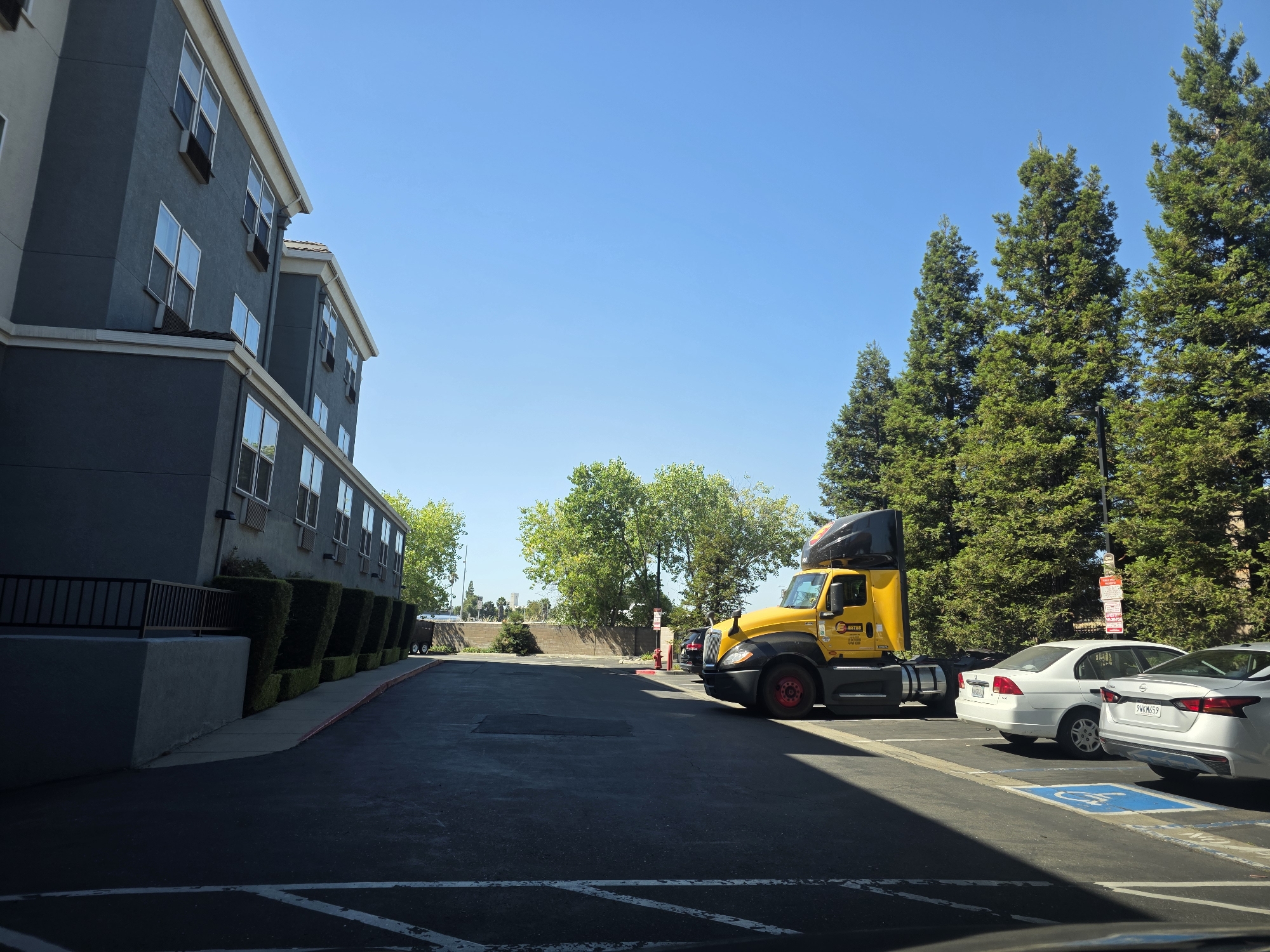 A bright, sunny day in West Sacramento, California. The image captures an outdoor scene with a large, multi-story building on the left, featuring a dark gray facade and numerous windows. A neatly trimmed hedge lines the side of the building. In the center of the frame, a bright yellow semi-truck is parked, its branding visible. To the right of the truck, two white cars are parked in designated spots, one with a handicapped parking symbol. Tall evergreen trees form a backdrop on the right side of the image. The sky is clear and blue, indicating fair weather. The sun casts shadows, suggesting it is mid-morning or early afternoon. The scene appears to be a parking lot adjacent to a commercial building. Visible text on the truck includes "ESTES" and "2520". One of the cars has a license plate that reads "PWKM659".