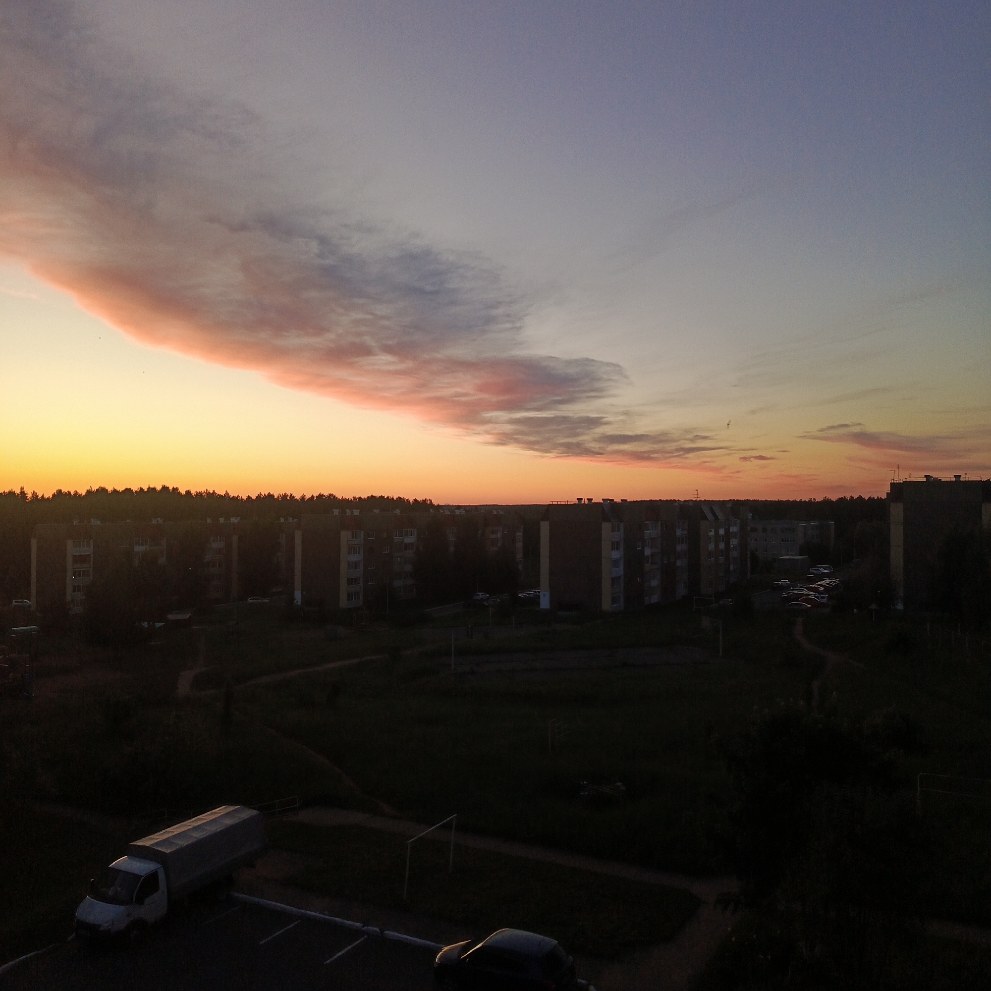 This elevated view captures a residential area at either sunrise or sunset, characterized by a dramatically colorful sky. The sky presents a vibrant gradient, transitioning from bright yellow and orange at the horizon, through shades of pink and purple, to a deep blue at the top. A distinct, elongated cloud band, colored in warm pinks and oranges, stretches across the upper left portion of the sky.
Below the colorful horizon, a dense line of trees forms a dark silhouette, bordering multiple multi-story apartment buildings. These residential structures, indicative of Soviet-era architecture, are partially illuminated by the low sun's warm light or cast in deep shadow, blending into the darkening landscape. In the foreground, expansive green spaces, likely fields or unkempt parks, are largely in shadow, with faint paths winding through them.
A white utility truck with a grey tarped bed is prominently parked in the bottom left, its top surfaces catching the golden light of the sun. Another dark-colored car is partially visible in the bottom right, similarly touched by the low sunlight. Several other cars are parked near the apartment buildings. No people are visible in the scene. The image depicts a tranquil transition of day in an urban environment that borders natural landscapes, consistent with a location such as Марковский, Russia. No visible text is present.