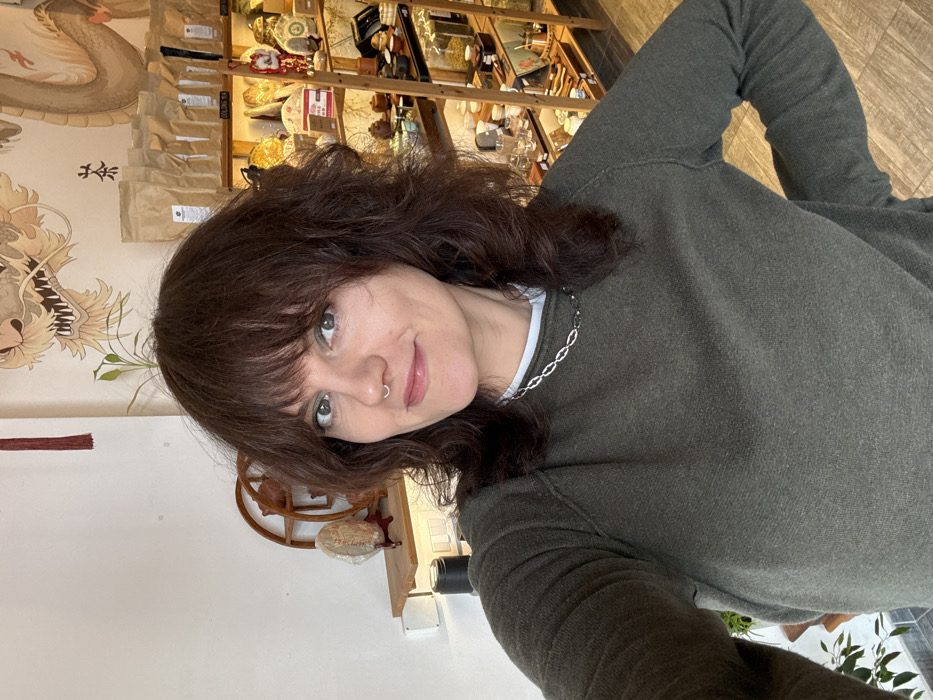 A female subject is captured in a medium shot selfie, smiling faintly and looking directly at the camera. She possesses shoulder-length dark brown hair with bangs, light skin, green eyes accented with green eyeshadow, and a silver septum ring. She wears a dark olive green long-sleeved sweater over a white undershirt, complemented by a silver chain necklace. Her right arm is bent, with a hand partially visible near her hip. The indoor background indicates a commercial establishment, likely a tea shop or an Asian cultural goods store. A large mural depicting a golden dragon with red details is prominent on the wall behind her. To the right, a multi-tiered wooden shelving unit displays numerous items including bags labeled "OOLONG TEA," ceramic tea sets, tea tins, and glass containers with what appear to be dried herbs or grains. To the left, a smaller wooden shelf holds a round, flat object on a stand, consistent with a tea cake, and a decorative wooden item. A small green plant is visible on the wall above this shelf. The specific location within the city of Budva, Montenegro, cannot be ascertained from the visual evidence presented.
