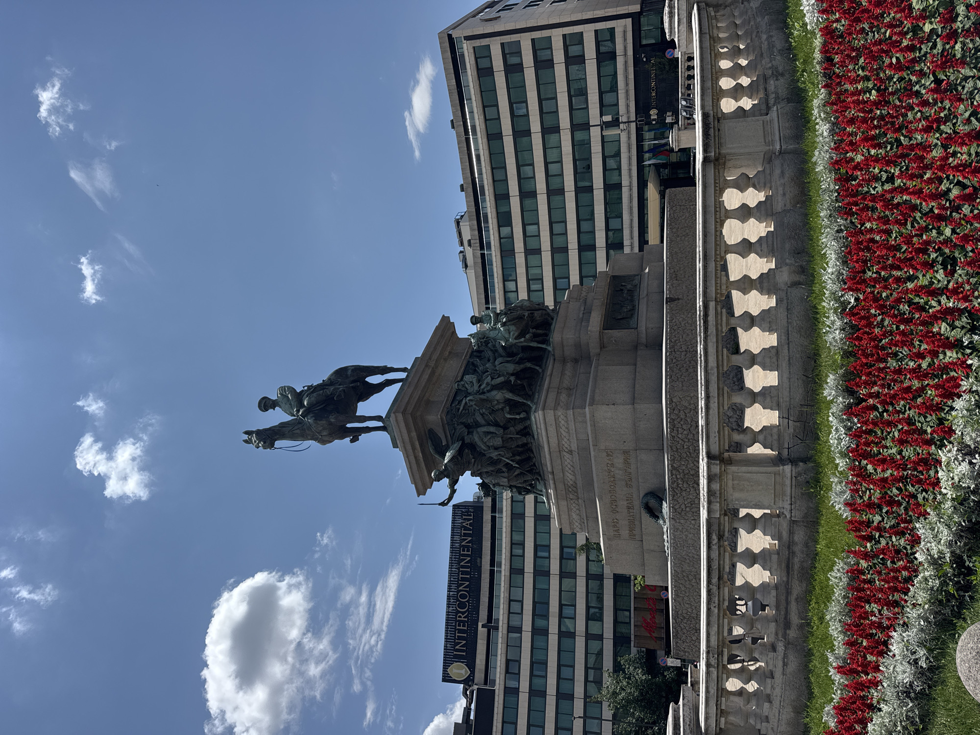 The image displays a clear, sunlit view of the Monument to the Tsar Liberator in Sofia, Bulgaria. Centrally positioned, a large bronze equestrian statue depicts a uniformed male figure on horseback, facing slightly right. This statue rests on a tall, multi-tiered stone pedestal adorned with several bronze bas-reliefs illustrating historical scenes with numerous figures, including soldiers and civilians; one relief figure is shown holding a sword aloft. An inscription in Cyrillic, "ЦАРЬ ОСВОБОДИТЕЛЬЮ ПРИЗНАТЕЛНА БЪЛГАРИЯ" (Tsar Liberator, Grateful Bulgaria), is visible on the pedestal, with a wreath placed at its base.
In the background, to the left of the monument, stands a multi-story modern building with the "INTERCONTINENTAL" logo prominently displayed in large, illuminated letters across its upper section. The building's facade consists of light-colored panels and multiple horizontal rows of dark-framed windows. A similar building is partially visible to the right.
The sky overhead is predominantly clear blue with scattered, sparse white cumulus clouds. In the immediate foreground, a low, curved stone balustrade with decorative balusters separates the viewer from a manicured garden bed. This garden features organized rows of vibrant red flowering plants, possibly Salvia splendens, interspersed with rows of silver-leafed foliage and green grass. The scene is brightly illuminated by direct sunlight, casting visible shadows.