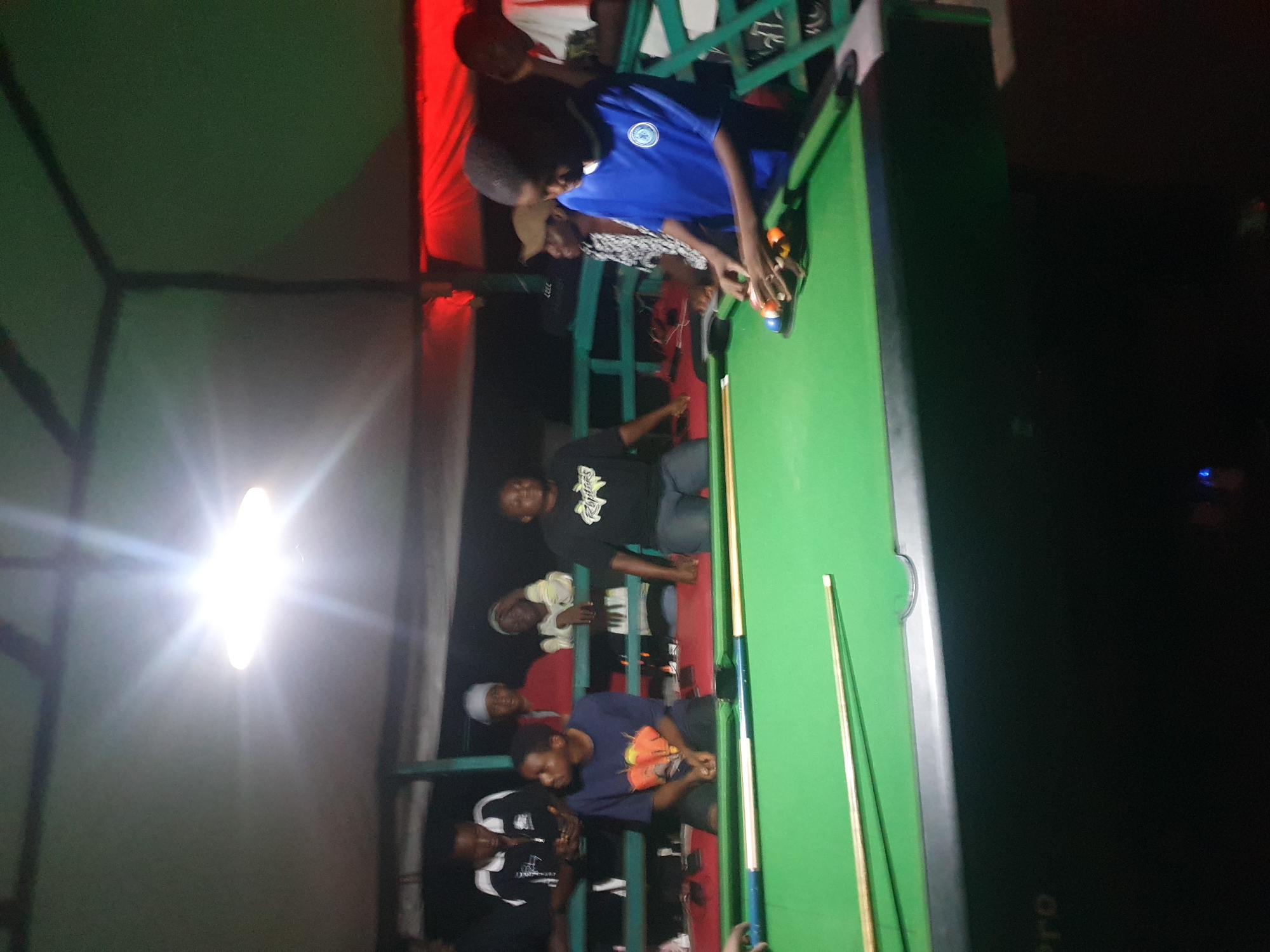 The image shows an indoor pool hall, likely in Lokoja, Nigeria, at night. A bright overhead light illuminates the scene. Several young men are present, some standing and watching, while others are seated or engaged with a pool table. In the foreground, a young man in a blue shirt is actively playing pool, lining up a shot with several colored balls on the green felt table. Two pool cues are visible on the table. The overall environment appears casual and dimly lit, with a red-lit area visible in the background.