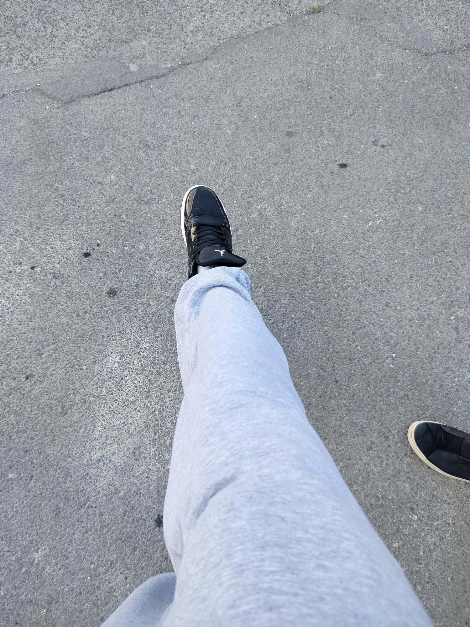 The image presents a first-person perspective of a person standing on a concrete surface in Yerevan, Armenia. The individual is wearing grey sweatpants and two different types of shoes. On the left, there is a black and white Air Jordan sneaker. On the right, there is a black slip-on shoe with a white sole. The concrete surface appears to be a sidewalk or street, showing typical wear and tear. The lighting suggests a daytime shot with ample natural light. There are no other people visible in the frame.