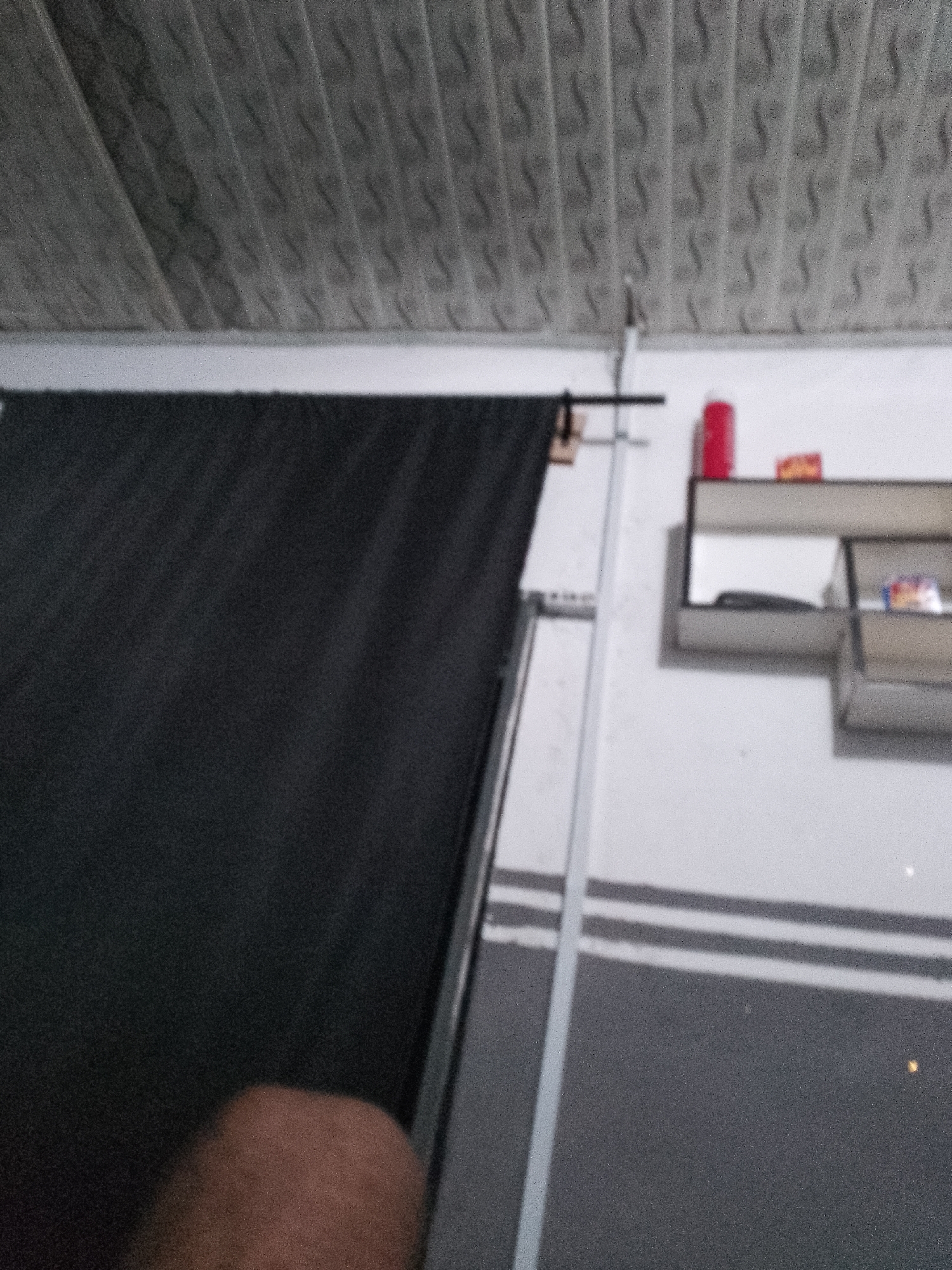 In an indoor setting, the scene depicts a wall with horizontal grey stripes, a portion of a black curtain, and a small shelf mounted on the wall. The shelf holds a red cylindrical object, a rectangular object, and a box with colorful packaging. The ceiling appears to be made of textured panels. A person's hand is visible in the lower left corner of the frame. The overall image is blurry.