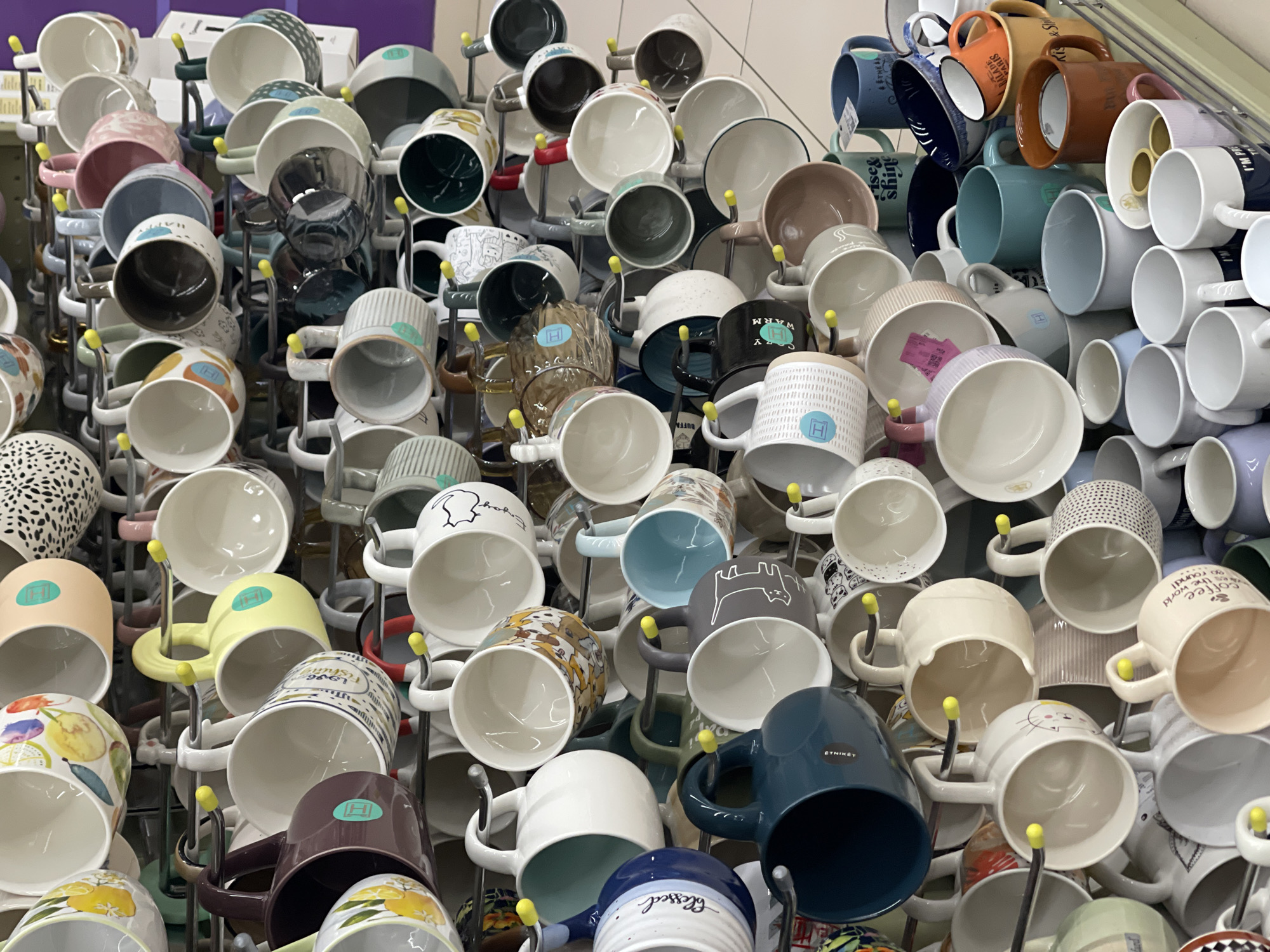 A dense display of mugs is organized on metal racks with yellow-tipped pegs. The mugs vary in color, pattern, and design, ranging from solid colors like white, blue, brown, and teal, to patterns such as polka dots, stripes, and various graphic illustrations, some containing words like "blessed," "coffee," and "Paris." Some mugs feature fruit patterns, others have animal illustrations. The handles of the mugs are visible and also vary in color. The mugs are positioned with their interiors facing upward. The location appears to be a retail environment.