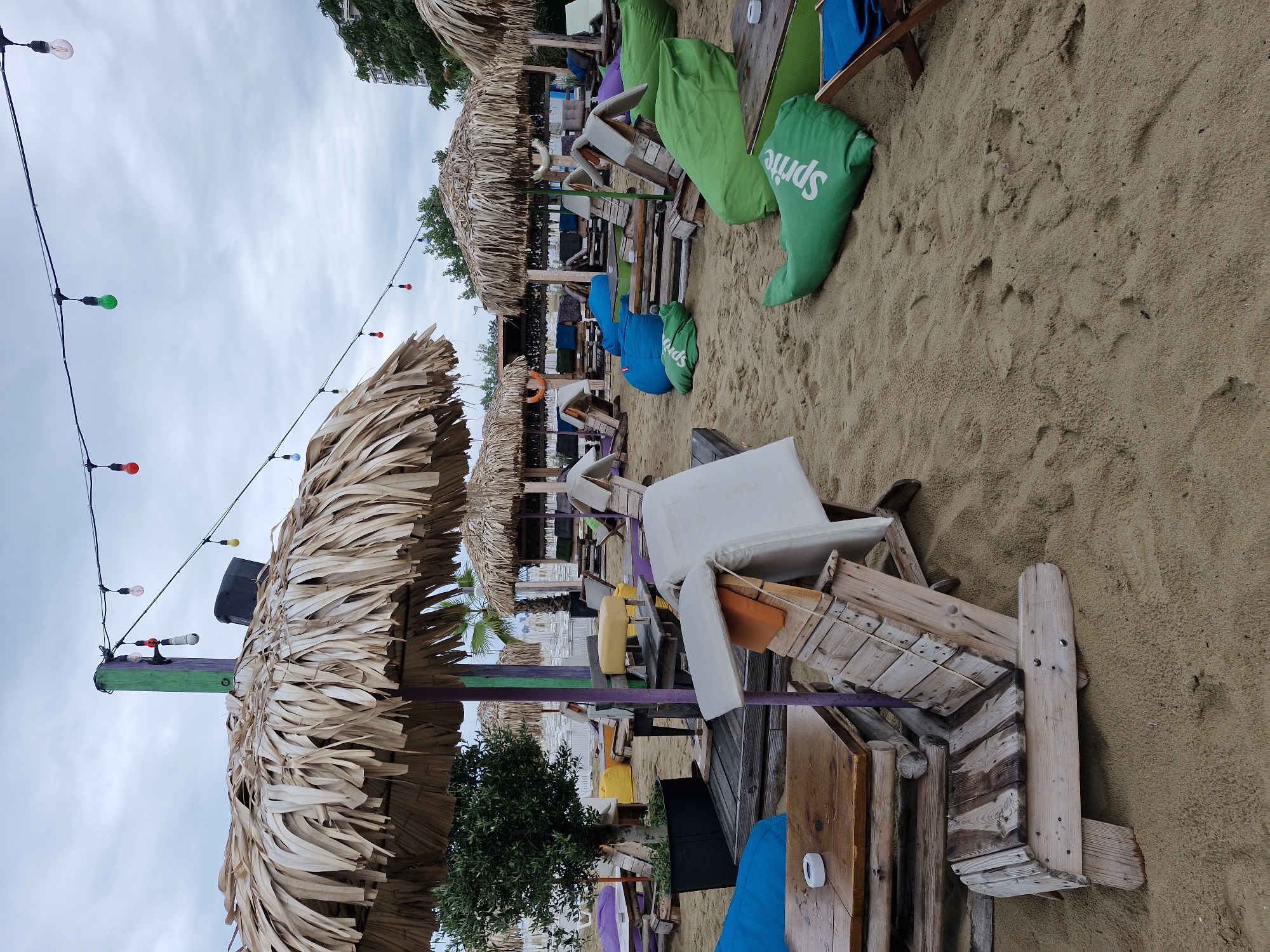 The image depicts a beachside seating area in Nesebar, Bulgaria. The scene is set on a sandy beach with several rows of seating arrangements under thatched umbrellas. Various seating options are visible, including wooden chairs, beanbag chairs in blue and green, and cushioned benches. Tables are scattered throughout the area, some made of wood and others with green tops. String lights with colored bulbs are suspended overhead. In the background, more beach umbrellas and structures are visible, along with some buildings and trees. The sky is overcast. There are no people clearly visible in the image.
