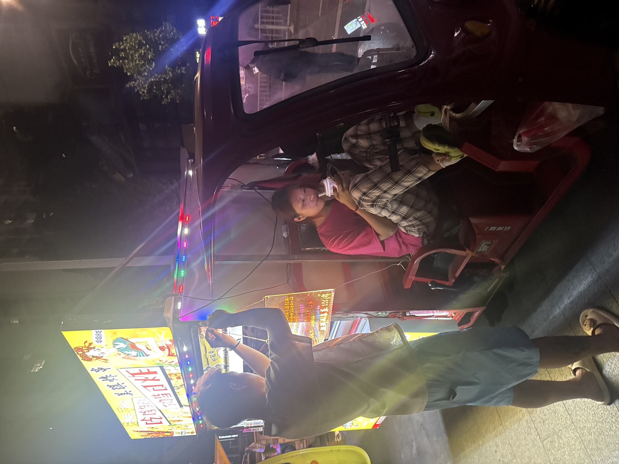 At night in Yuzhong District, China, a man stands facing a lit sign in a brightly lit street food stall, seemingly fixing something. A woman sits in the vehicle of the stall behind him, looking at her phone.