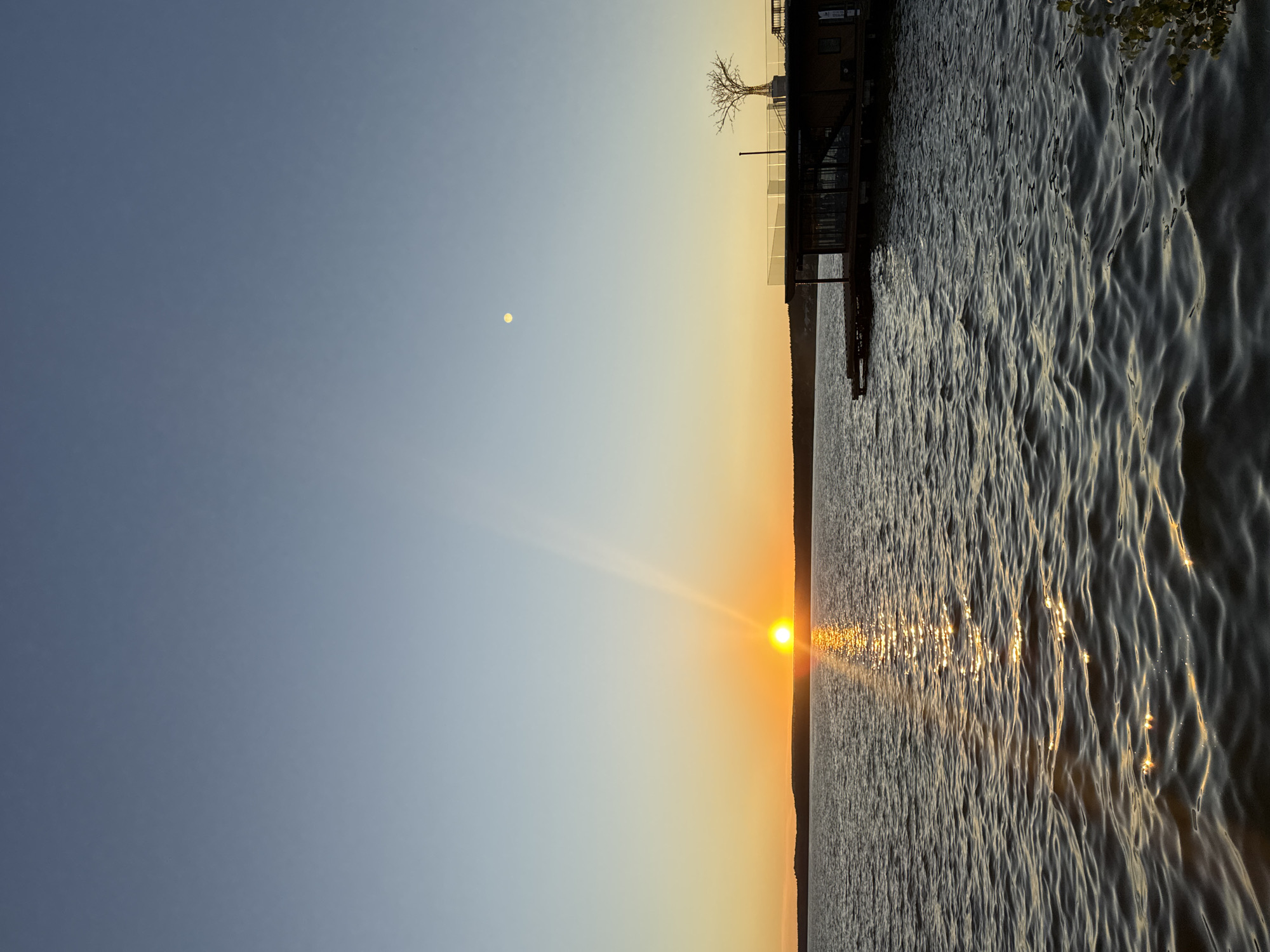 The image depicts a serene sunset over the Votkinsk Reservoir in Votkinsk, Russia. The sun, a bright orange orb, is low on the horizon, casting a shimmering golden path across the rippling water. A distant shoreline with a dark silhouette of trees stretches across the frame. To the right, a modern, multi-level structure resembling a restaurant or lodge is situated on a wooden pier extending into the water. The structure has large windows, and a railing encircles the upper level where a leafless, illuminated tree stands as a decorative element. In the clear, pale blue sky, a small, pale circle, possibly the moon, is visible. The overall atmosphere is peaceful and tranquil, capturing the natural beauty of the location during twilight.