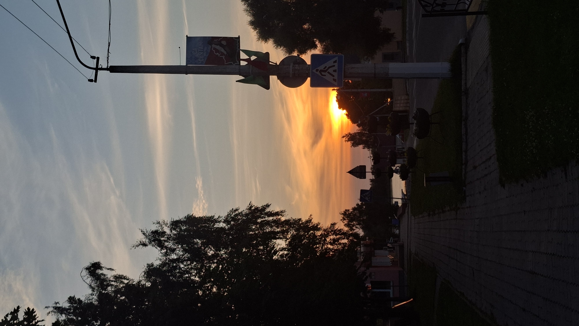 The image captures a sunset scene in the city of Myadzyel, Belarus. In the foreground, a brick-paved sidewalk runs along the left side of the frame, bordered by grass and some dark planters. A utility pole stands prominently on the right, adorned with several signs, including a blue pedestrian crossing sign and a banner with text. The sun is setting on the horizon, casting a warm orange and yellow glow across the sky, which is streaked with wispy clouds. Silhouetted trees frame the scene on the left and right. In the middle ground, a road is visible, and beyond that, a body of water reflects the sunset colors. Buildings are faintly visible on either side of the street. The overall impression is one of a tranquil evening in a Belarusian town.