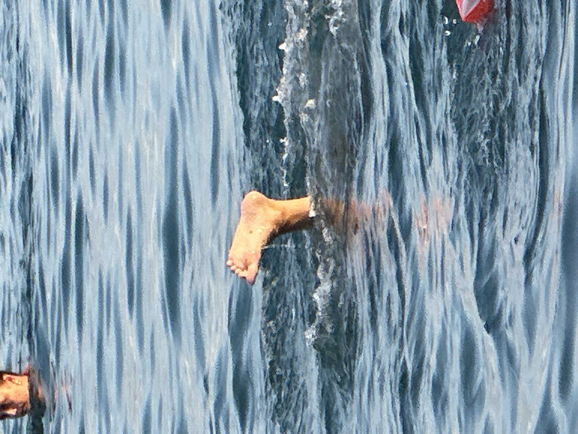 In the city of Sochi, Russia, an image captures a person swimming in the Black Sea. The upper portion of a person's head and shoulder is visible at the top left of the frame, indicating they are partially submerged. In the center of the image, a bare foot with visible toes emerges from the water, with droplets of water cascading down from it. The water is calm with gentle ripples, suggesting a peaceful environment. A red object, possibly a buoy or a float, is partially visible in the bottom right corner of the frame. The scene appears to be a leisure activity on a sunny day, with the water reflecting the sky and sunlight.