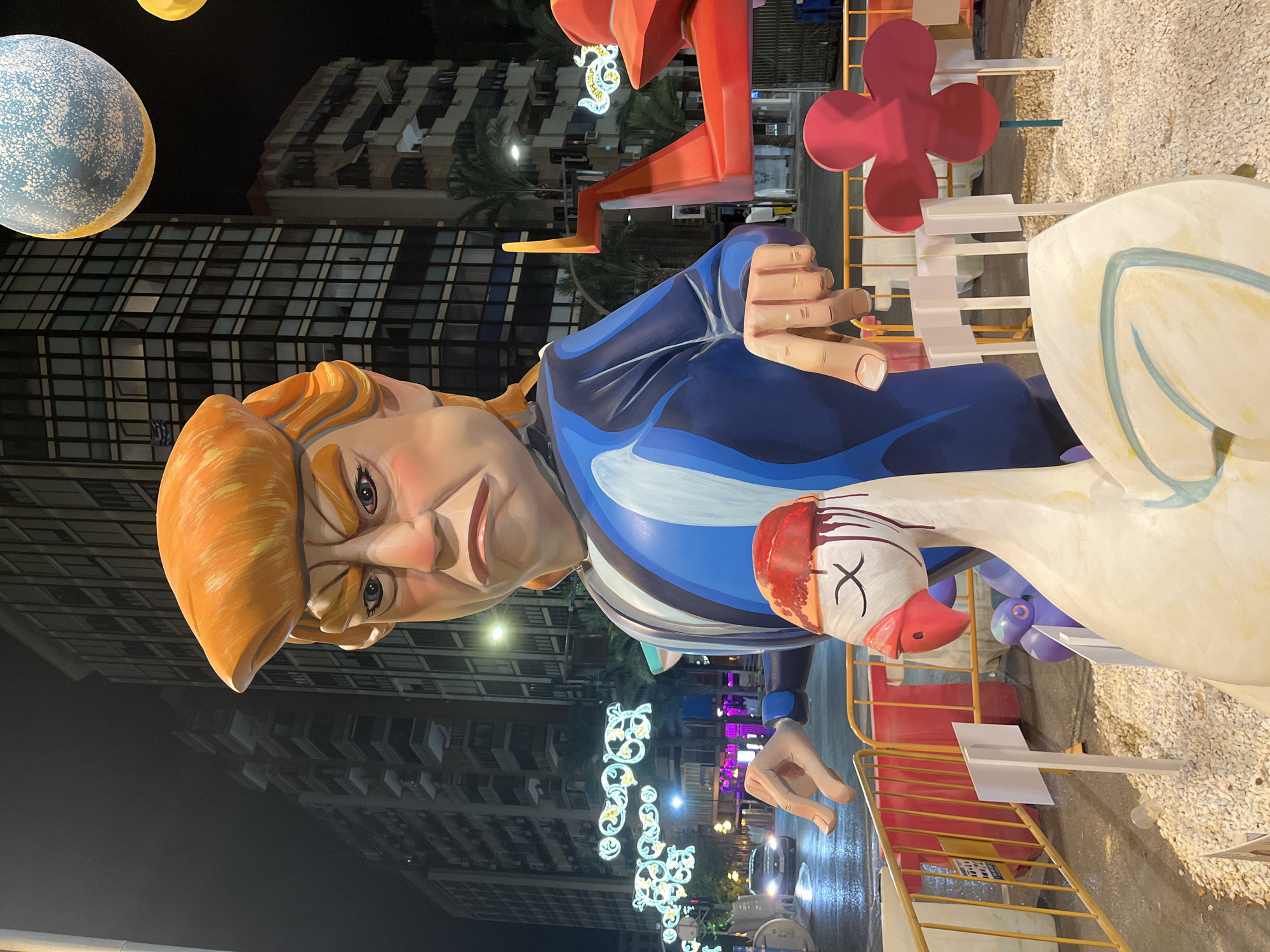 The image shows a large, cartoonish effigy of Donald Trump, dressed in a blue and striped suit, in Alicante, Spain. The effigy is part of a larger display, with other elements including a large white swan-like creature with a red head, and a red airplane-like structure. The scene is illuminated by streetlights and decorative lights, suggesting it is nighttime. In the background, tall buildings are visible. The effigy is positioned in a public space, with yellow barriers in the foreground. The overall scene depicts a festive or satirical art installation.