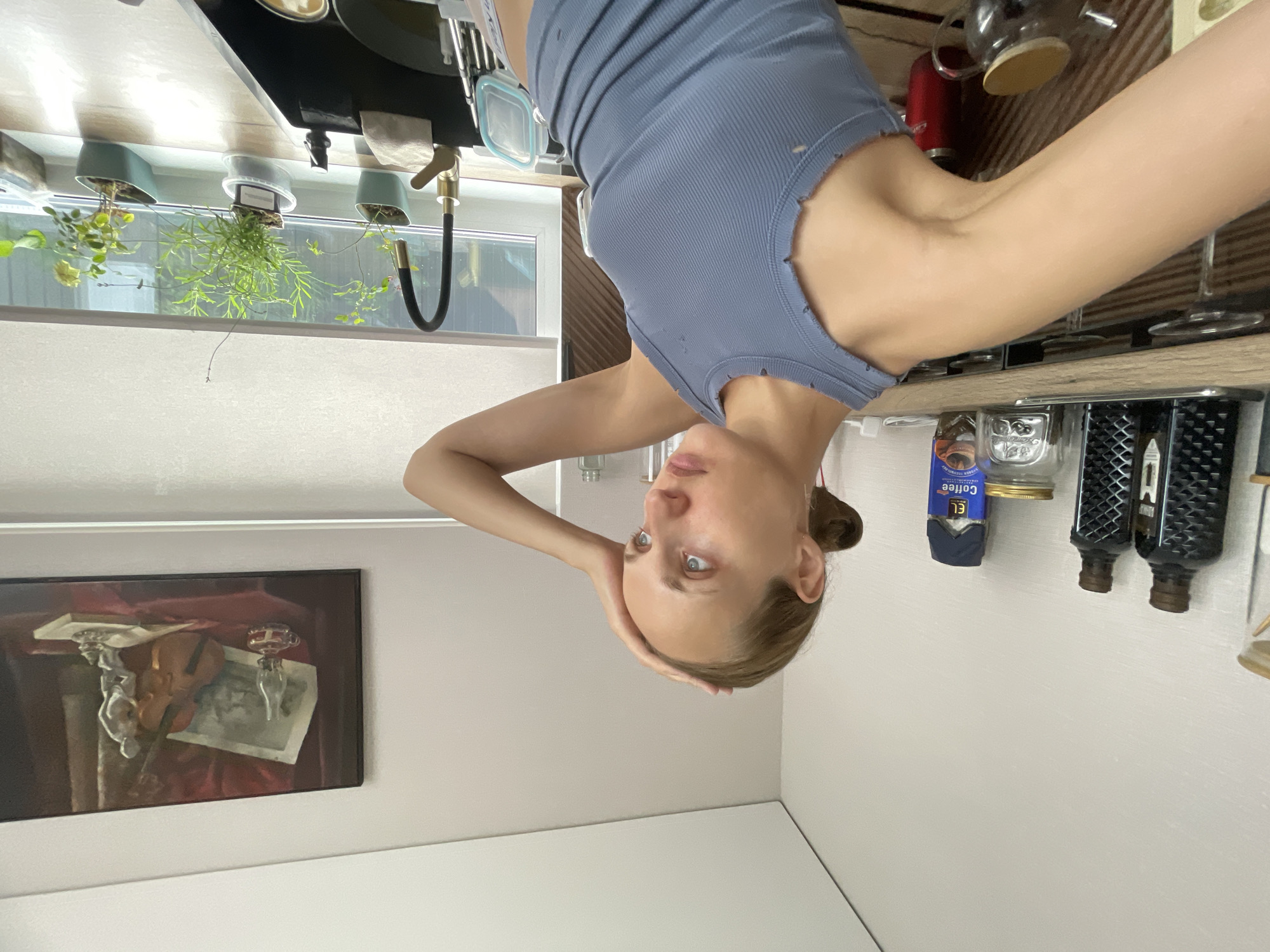 A young woman with blonde hair pulled back in a bun, wearing a blue ribbed tank top, stands in a kitchen. Her left hand is on her forehead, and her gaze is directed to the right, with a pensive expression. The kitchen counter is visible, holding several items including glass jars with coffee, dark bottles, and a red container. A painting featuring a violin, a lamp, and a statue hangs on the wall above the counter. A sink with a black basin and a gold faucet is in the foreground, with several potted plants on the windowsill behind it. A window covered by a roller blind is also present. The scene is lit by natural light. The location is identified as Podshivalovo, Russia.