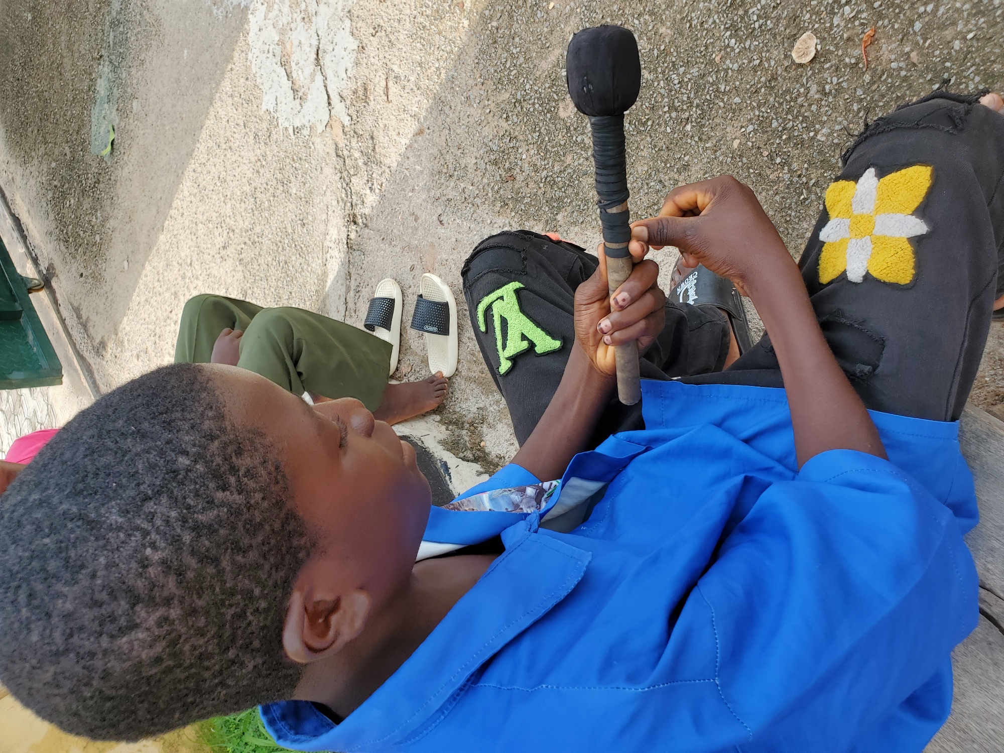 This image, taken in Mararaba, Nigeria, presents an overhead view of a young person reclining outdoors, with parts of two other individuals also visible. The primary subject, seen from the chest up and also showing their legs, is wearing a bright blue shirt or jacket and black pants. The black pants feature two distinct, colorful patches: a large, bright green letter "A" or similar symbol on the left thigh, and a checkerboard-patterned yellow and white flower-like design on the right thigh.
The main subject is holding a wooden stick-like object vertically with both hands. The top of the stick is covered with a black, bulbous fabric, resembling a makeshift microphone or a ceremonial staff, and the handle is wrapped in black tape or fabric. The person's gaze is directed slightly upwards and to the left, suggesting a moment of rest or contemplation.
To the left and slightly behind the main subject, another person's lower body is visible, clad in olive green pants and white slide sandals with black straps. The word "CROWN" is discernible on the strap of one of the sandals. A bare foot, likely belonging to a third individual, is partially seen near the sandals.
The setting appears to be an outdoor area with a rough, light-colored concrete or asphalt ground, showing some dirt and small debris. Sunlight suggests it is daytime, and shadows are visible on the ground. A green object, possibly a part of a metal gate or structure, is on the far left, and a wooden surface is visible on the far right. The overall scene depicts a casual, informal outdoor gathering or moment of relaxation.