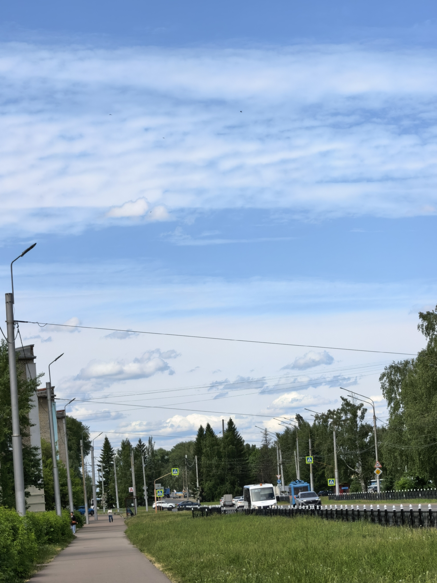 The image shows an urban street scene in Sterlitamak, Russia, on a bright, partly cloudy day. A paved sidewalk stretches along the left side of the frame, with several pedestrians walking along it. To the right of the sidewalk, a green grassy median separates it from the road. A black metal fence runs along the edge of the median, bordering the road. Various vehicles, including a white van and several cars, are visible on the road. Traffic signs, including pedestrian crossing signs, are placed along the street. Tall trees line both sides of the road, creating a green canopy in the background. Streetlights are also present along the road, supported by metal poles. The sky is blue with white clouds, taking up the majority of the upper portion of the image. The image suggests a typical summer day in an urban area.
