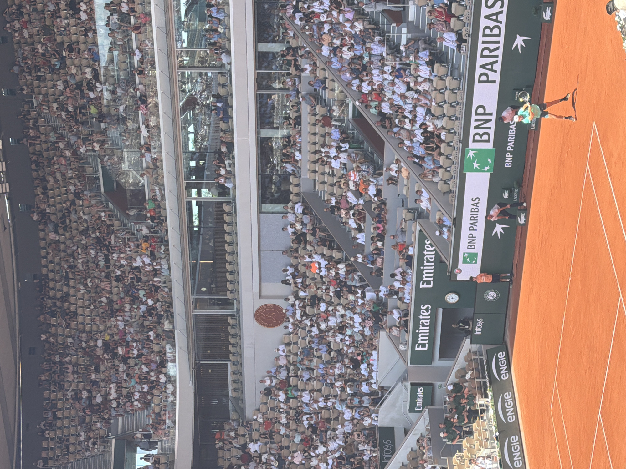 The image depicts a tennis match being played at Roland Garros in Paris, France. The court is clay, and a player is in the midst of a serve. The stands are filled with spectators. The stadium is surrounded by signs advertising Emirates, Infosys, BNP Paribas, Lacoste, and ENGIE.