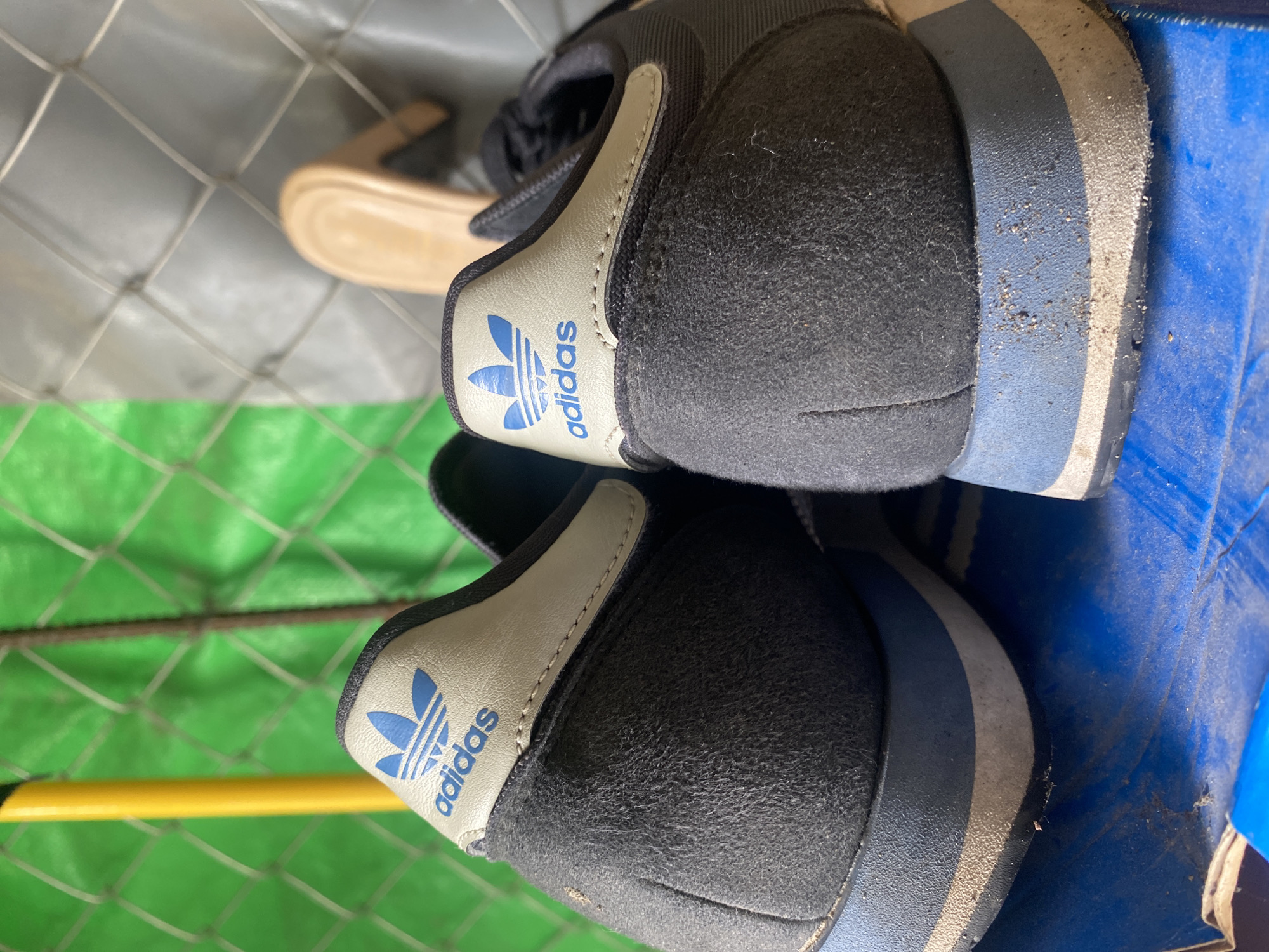 The image, taken in Davao City, Philippines, displays two pairs of shoes, with a primary focus on the rear view of two black Adidas sneakers. Each sneaker features a black, slightly fuzzy upper material, and a light grey/beige heel tab embossed with the blue Adidas trefoil logo and "adidas" text. The soles are distinctly multi-layered, showing sections of black, light blue, and grey/silver, with visible dust and signs of wear. One sneaker is positioned slightly higher and behind the other, both resting against a surface.
In the blurred mid-ground, a light brown shoe or sandal is partially visible. The background is characterized by a prominent green and grey/silver grid pattern, likely a fence or net, with a horizontal yellow bar running across the lower left section. To the right, a bright blue surface, appearing to be a box or storage container, serves as a support for the sneakers. The shoes appear to be stored or resting in a casual setting, possibly an outdoor or utility area, under bright daytime lighting. No specific activity is depicted beyond the static placement of the shoes.