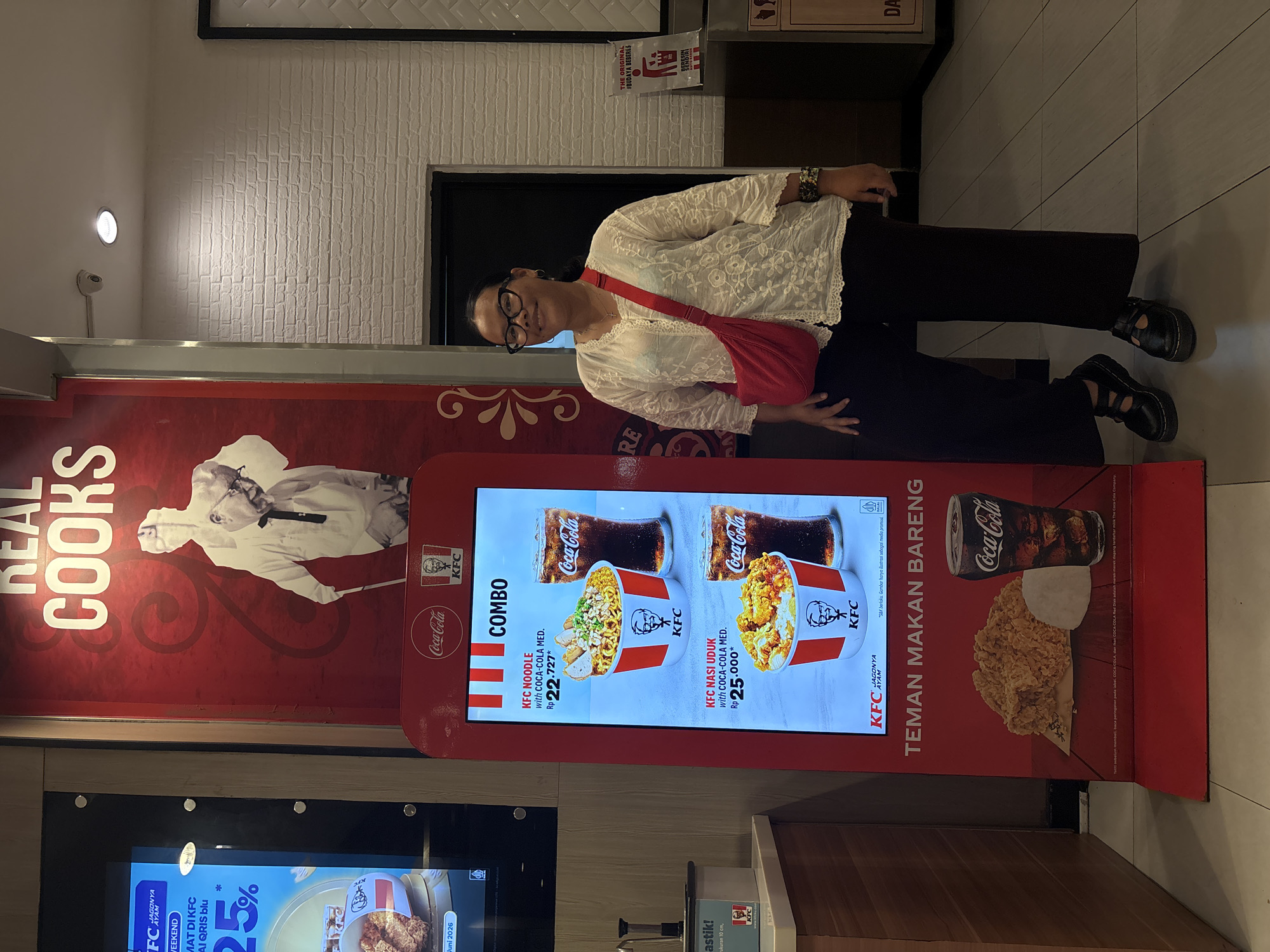 A woman stands in front of a KFC digital menu board in Kuta Selatan, Indonesia. The menu board displays two combo meal options: KFC Noodle with Coca-Cola Medium for Rp 22,727 and KFC Nasi Uduk with Coca-Cola Medium for Rp 25,000. The woman is wearing glasses, a white lace top, and dark pants, and has a red crossbody bag. She is smiling at the camera. The KFC restaurant has a red and white color scheme, with a brick wall in the background. A trash can with a "THE ORIGINAL #BUDAYA BERES" sign is visible to the right of the woman.
