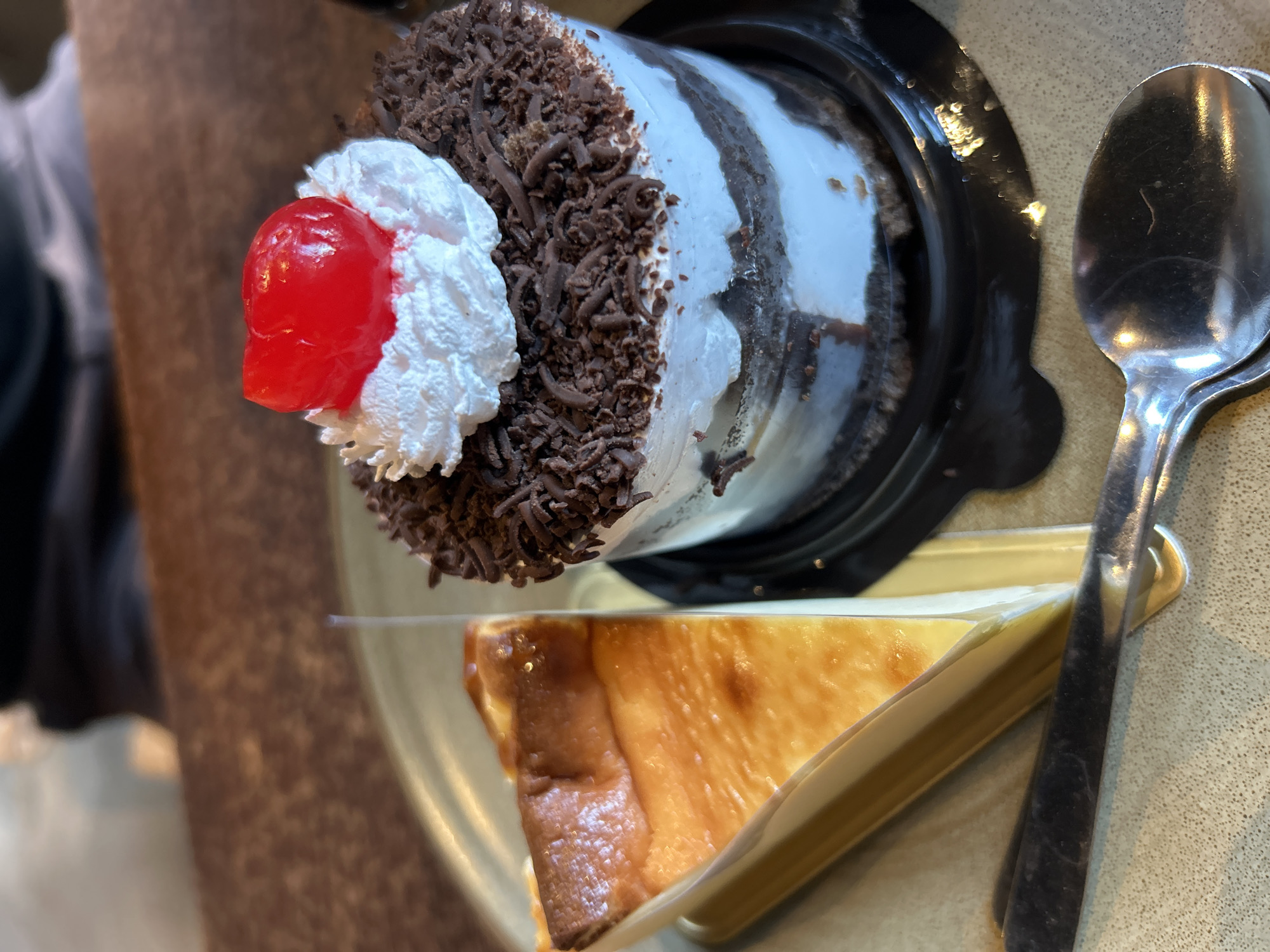 Two desserts are presented on a beige plate. To the left is a triangular slice of cheesecake with a golden-brown, slightly browned top and a pale yellow interior. The cheesecake is served on a golden paper liner. To the right is a cylindrical chocolate cake layered with white frosting and topped with whipped cream and a red cherry. The cake is adorned with chocolate shavings. A silver spoon rests on the plate in the foreground, partially overlapping the cheesecake. The background is blurred, showing a brown textured surface and a person wearing a dark shirt. The scene appears to be an indoor setting, possibly a cafe or restaurant, in the city of Sukawati, Indonesia, given the context provided.