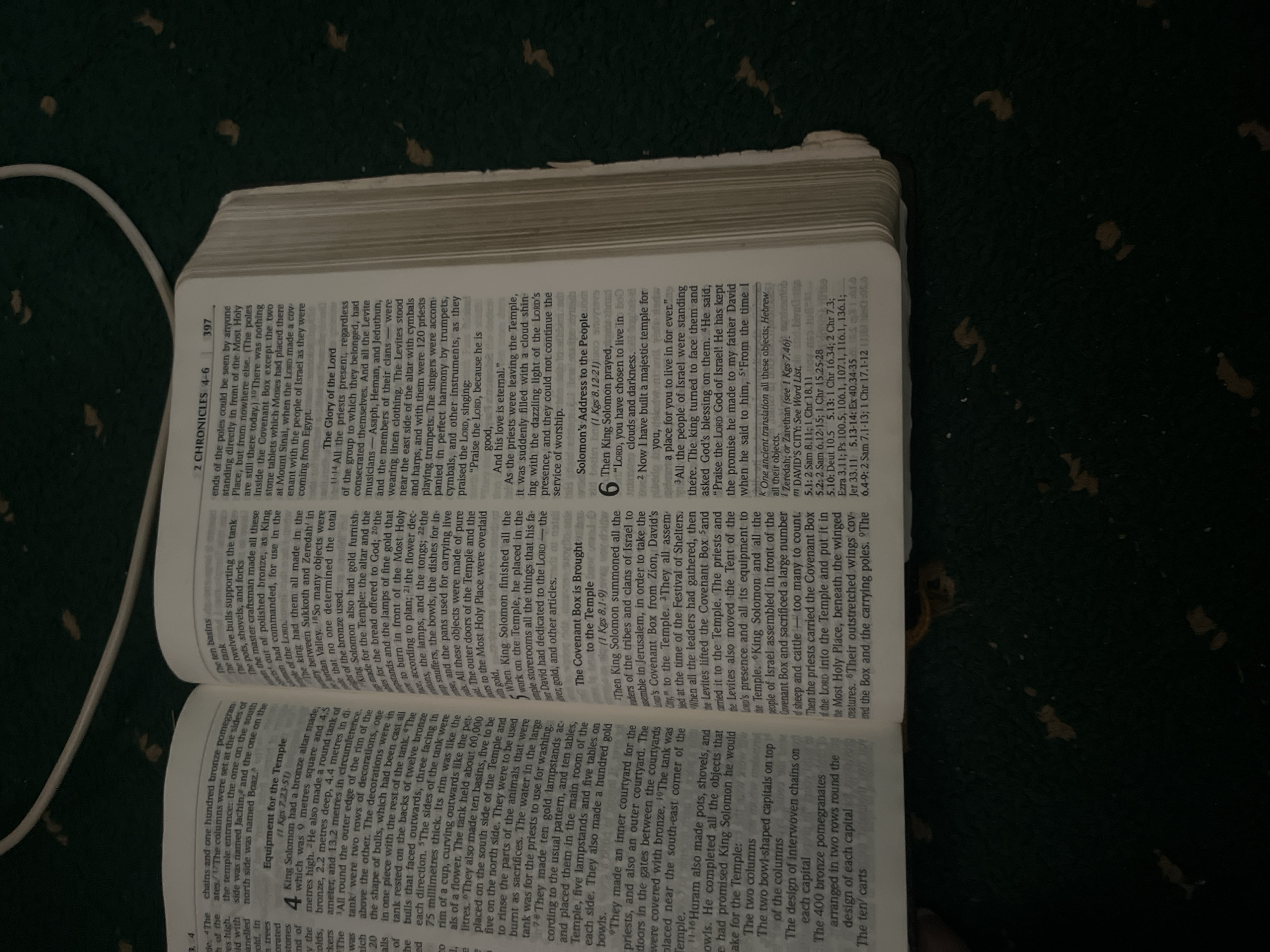 The image shows an open bible, with text on both pages. The left page contains text about the equipment for the temple, including details about bronze altars, pillars, and basins. The right page contains text from 2 Chronicles 4-6, discussing the glory of the Lord, Solomon's address to the people, and the Ark of the Covenant. The bible is lying on a dark, textured surface, possibly a carpet, with scattered light-colored debris, which could be dust or small pieces of paper. A white cord, likely from a lamp or charger, is visible in the top portion of the image. The lighting suggests it is indoors, possibly in a room with ambient or artificial light. There are no people or animals present in the image. The city of Jauro Yinu, Nigeria is not depicted or referenced in the image.