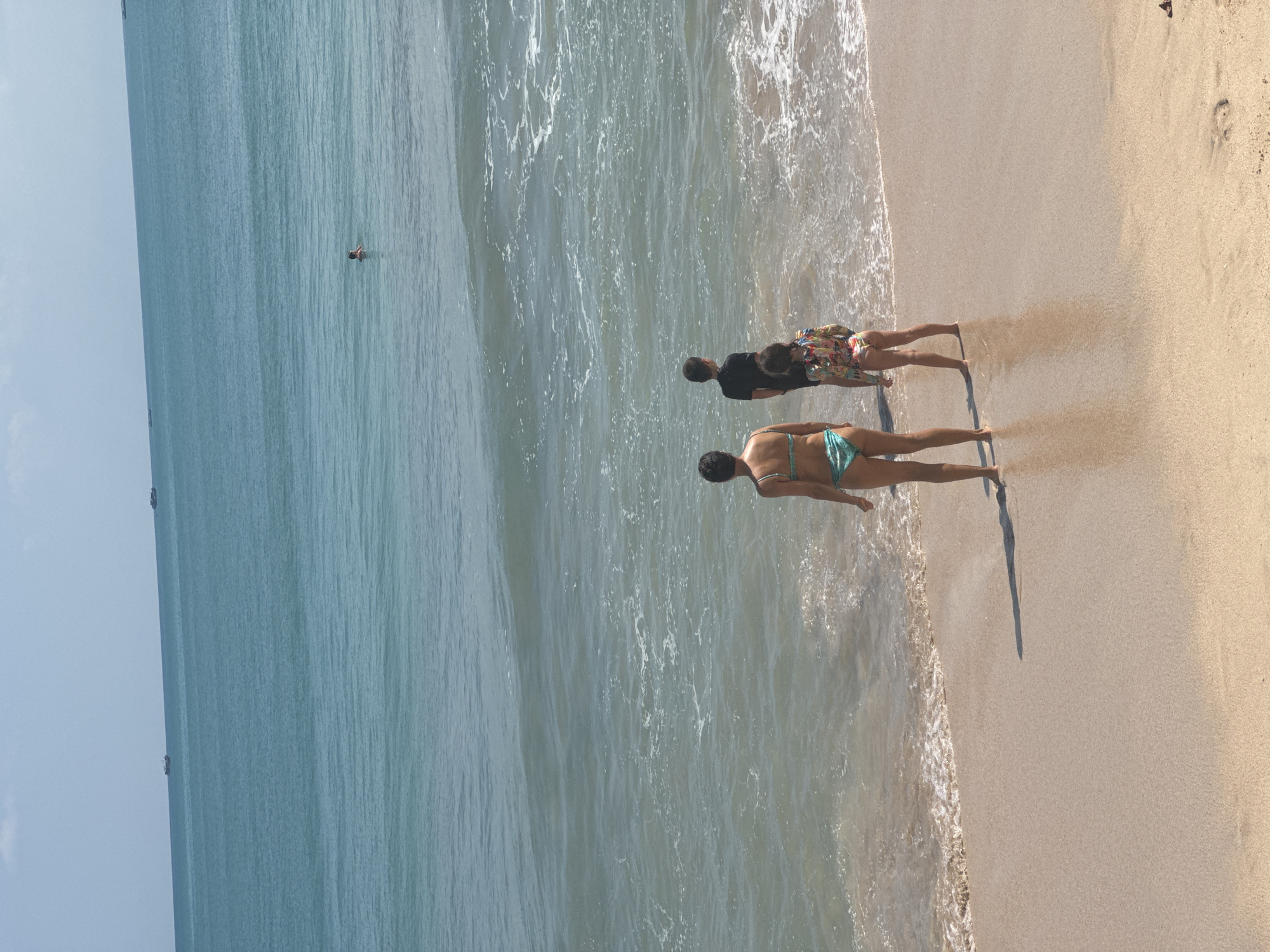 The image depicts a vibrant beach scene in what appears to be late morning or early afternoon, characterized by bright sunlight and clear skies. The perspective is from slightly above, looking down at the shoreline.
Three individuals are standing near the water's edge on a sandy beach. Closest to the viewer is a woman wearing a light blue two-piece bikini. To her right, slightly further back from the camera, stands a person in a black t-shirt and colorful patterned shorts. Adjacent to them, another individual is seen wearing a long-sleeved, brightly colored patterned top and matching shorts. All three are standing in the shallow, foamy water where gentle waves break. Their elongated shadows are cast clearly onto the wet sand, stretching towards the left of the frame.
Further out in the ocean, a solitary person is visible, seemingly floating or swimming. The ocean water is a clear blue-green, transitioning to a deeper blue in the distance. Several small boats are faintly visible on the horizon, indicating the expanse of the sea. The sandy shore is light brown and appears smooth, with some ripples from the receding waves.
The overall scene captures a relaxed and leisurely moment at a sunny, tropical beach, consistent with the location context of Kuta Selatan, Indonesia. There is no visible text in the image.