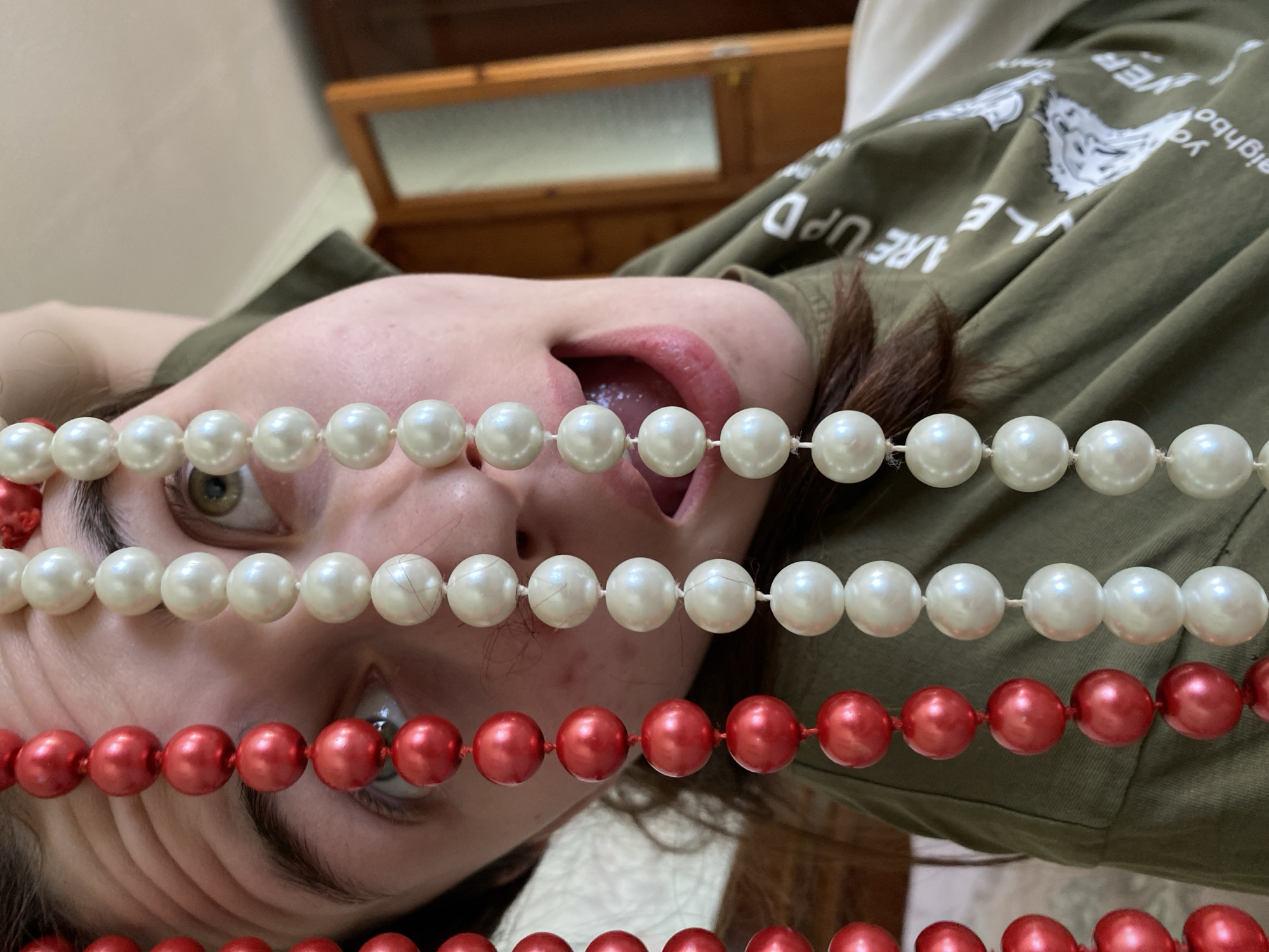 A person is lying down, facing the camera at an inverted angle. The person is wearing a green shirt with white printed graphics. The individual's mouth is open, revealing part of the tongue. Strands of beads are held in front of the face, consisting of two rows of white pearl-like beads and two rows of red beads. Part of a wooden cabinet is visible in the background. The image does not include any information or features that can reliably indicate it was taken in Votkinsk, Russia.