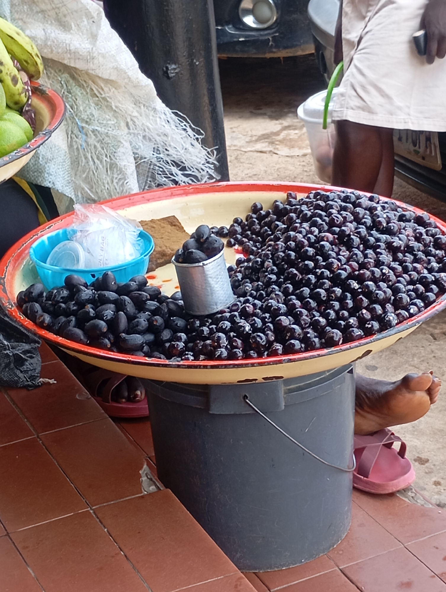 A close-up, eye-level shot captures an outdoor vendor's stall in Jos, Nigeria, displaying a large quantity of dark, oval-shaped fruits, likely African black olives, arranged on a round, shallow tray. The tray, with a red and yellow rim, is supported by a dark gray plastic bucket. To the left of the tray, a small blue bowl contains white plastic bags, and next to it, a small metal cup is filled with the same dark fruits. The fruits are abundant, filling the majority of the tray, with some spilling over. A person's bare foot, wearing a pink sandal, is visible resting on top of the bucket on the right side of the image. The background shows elements of an urban setting, including a vehicle's headlight and bumper on the left, and a person's lower legs and fabric of their clothing on the right, suggesting a busy street or market environment. The ground is a mix of reddish-brown tiles and concrete.