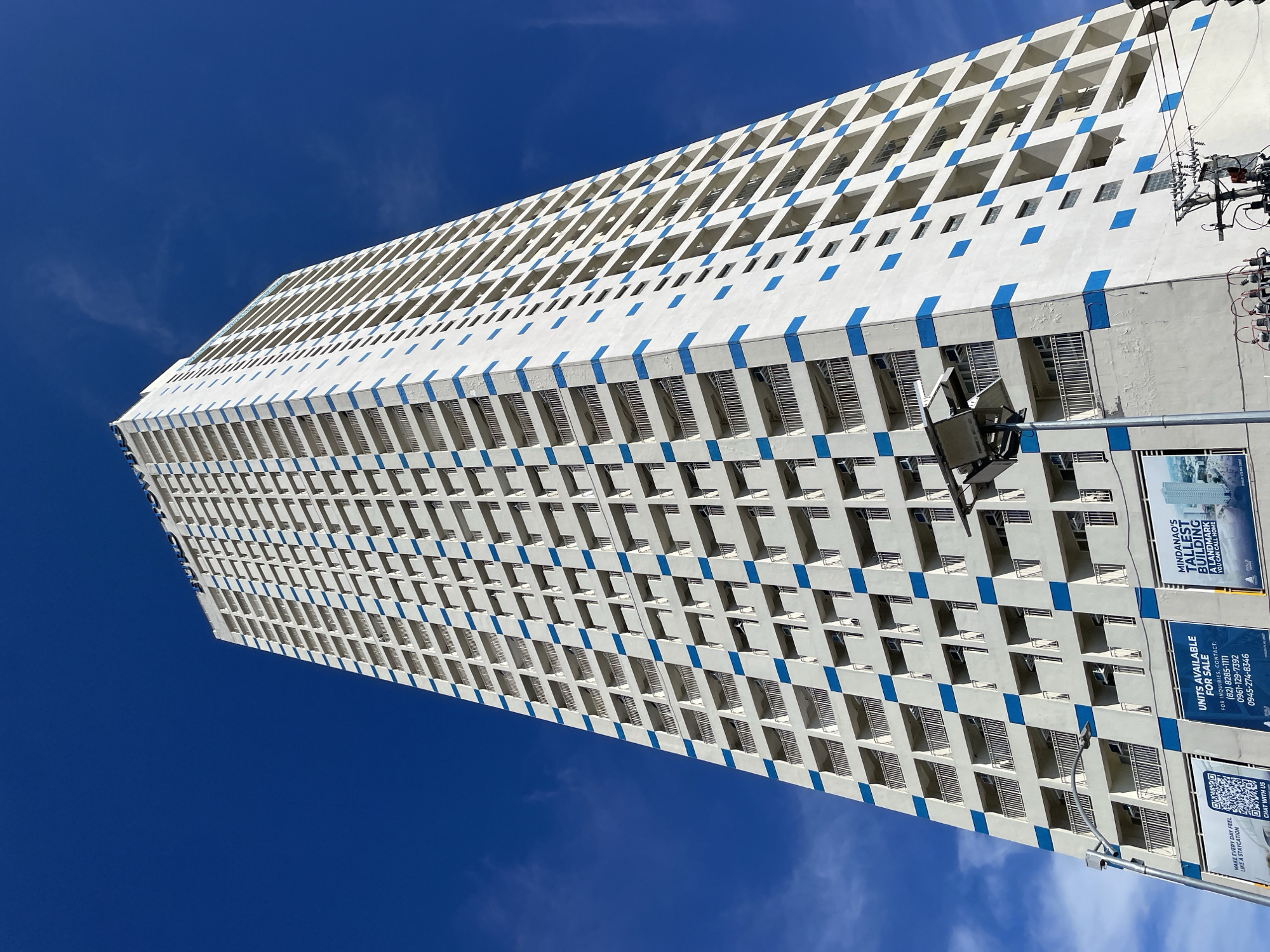 The image depicts a very tall, white, multi-story building with a pattern of blue squares on its exterior, set against a clear blue sky with scattered white clouds. The building appears to be a residential or commercial tower, with numerous balconies and windows visible. The perspective is from a low angle, emphasizing the height of the structure. On the lower portion of the building, there are several large advertisements or signs. One sign, in the bottom left, features a QR code and the text "MAKE EVERY DAY FEEL LIKE A STAYCATION. CHAT WITH US." Another sign, in the center, reads "UNITS AVAILABLE FOR SALE" with contact information, including phone numbers. A third sign on the right highlights "MINDANAO'S TALLEST BUILDING" and states "A LANDMARK YOU CAN CALL HOME." These advertisements suggest that the building is located in Davao City, Philippines, as indicated by the mention of Mindanao. There are also electrical wires and a street lamp in the foreground, indicating an urban setting. No people are visible in the image.