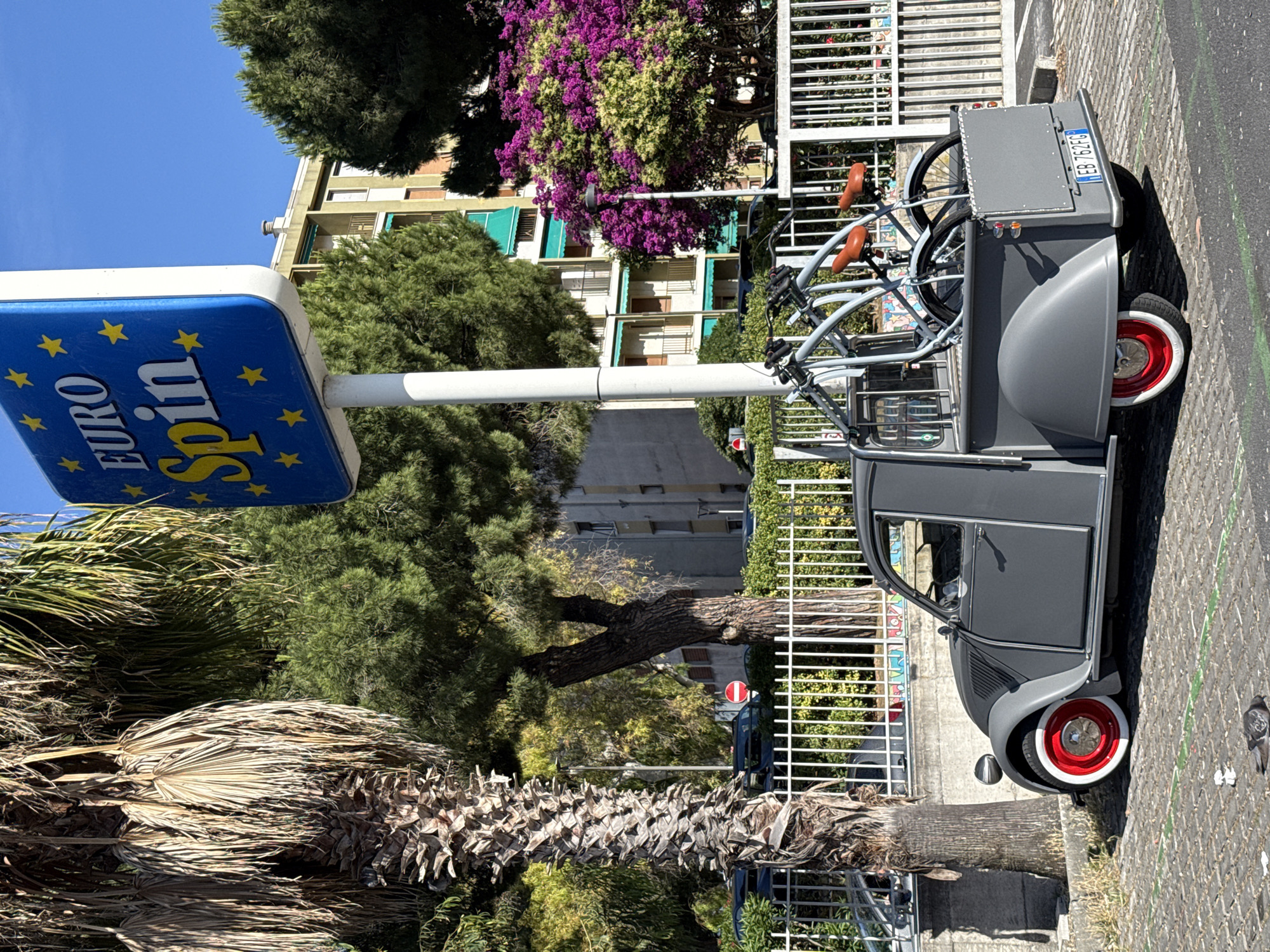 In Vallecrosia al Mare, Italy, a vintage grey Citroën 2CV pickup truck is parked on a cobblestone street. The truck has a customized rear bed fitted with a rack carrying two bicycles. The vehicles' wheels have white walls and red hubs. A tall palm tree with desiccated fronds stands to the left of the truck, casting a shadow. Behind the truck and fence, a building with green awnings is visible. A blue sign with yellow stars and the text "EURO Spin" is mounted on a pole to the right of the palm tree. The scene is under a clear blue sky on a sunny day.