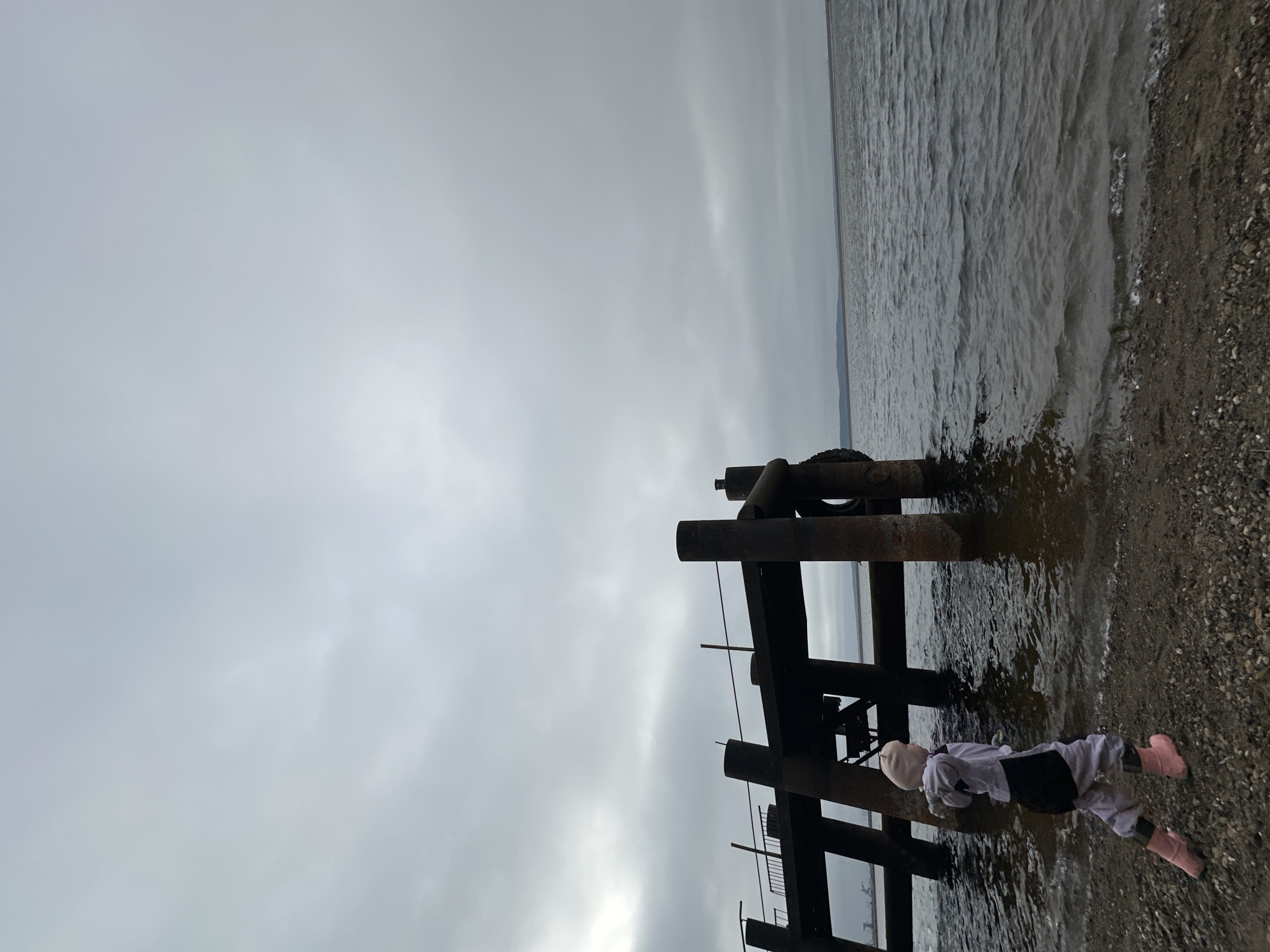 This image captures an overcast scene at the edge of a body of water in Chaikovsky, Russia. The sky is a uniform light grey, suggesting a heavily clouded day. To the right, a pebbly shore meets choppy, dark-grey water, which extends to the horizon.
A robust, weathered structure, possibly a pier or dock made of dark wood or metal beams, is centrally positioned, extending from the water onto the shore. A black tire is visible, attached to one of the structure's upright posts.
In the lower right, a small child is seen standing or wading in the shallow water next to the shore, partially obscured by the structure. The child is dressed in light-colored clothing, including a white top, grey pants, and a dark vest or jacket, and notably wears bright pink rain boots. Their back is turned to the viewer, facing towards the water and the structure. The mood is calm and muted due to the grey weather. No visible text is present.