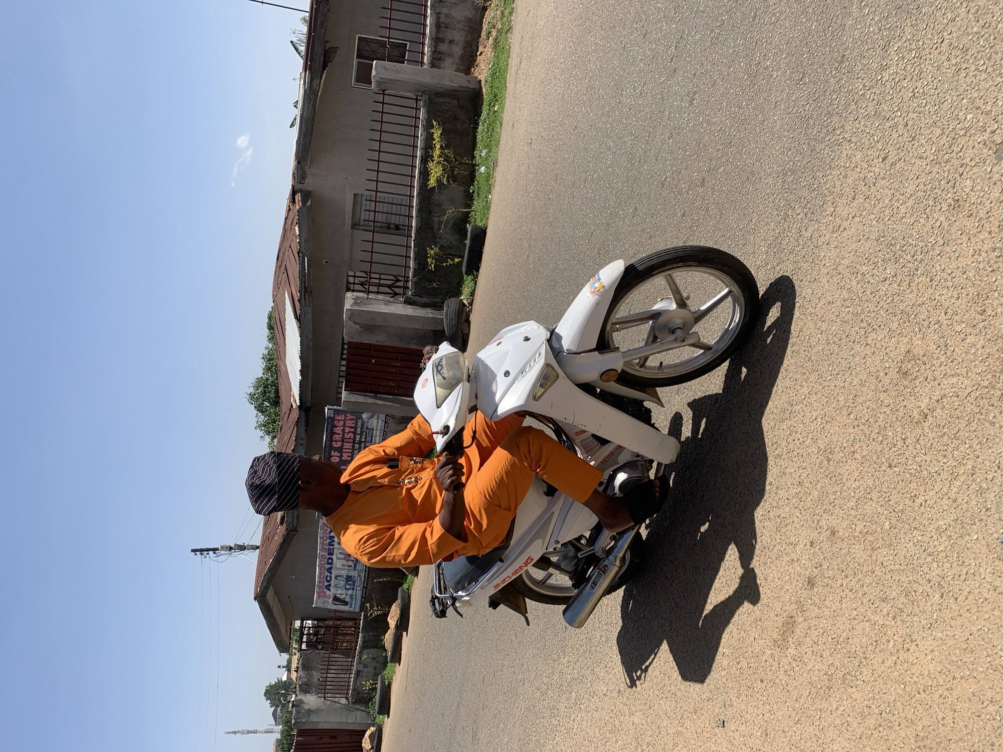 A man wearing an orange garment and a black and white striped cap sits astride a white Jincheng motorcycle. The motorcycle is parked on a paved road in front of a building. A sign reading "Precious Stone Academy" and "School of Grace Ministry" is visible on the building. The location appears to be in Jos, Nigeria.