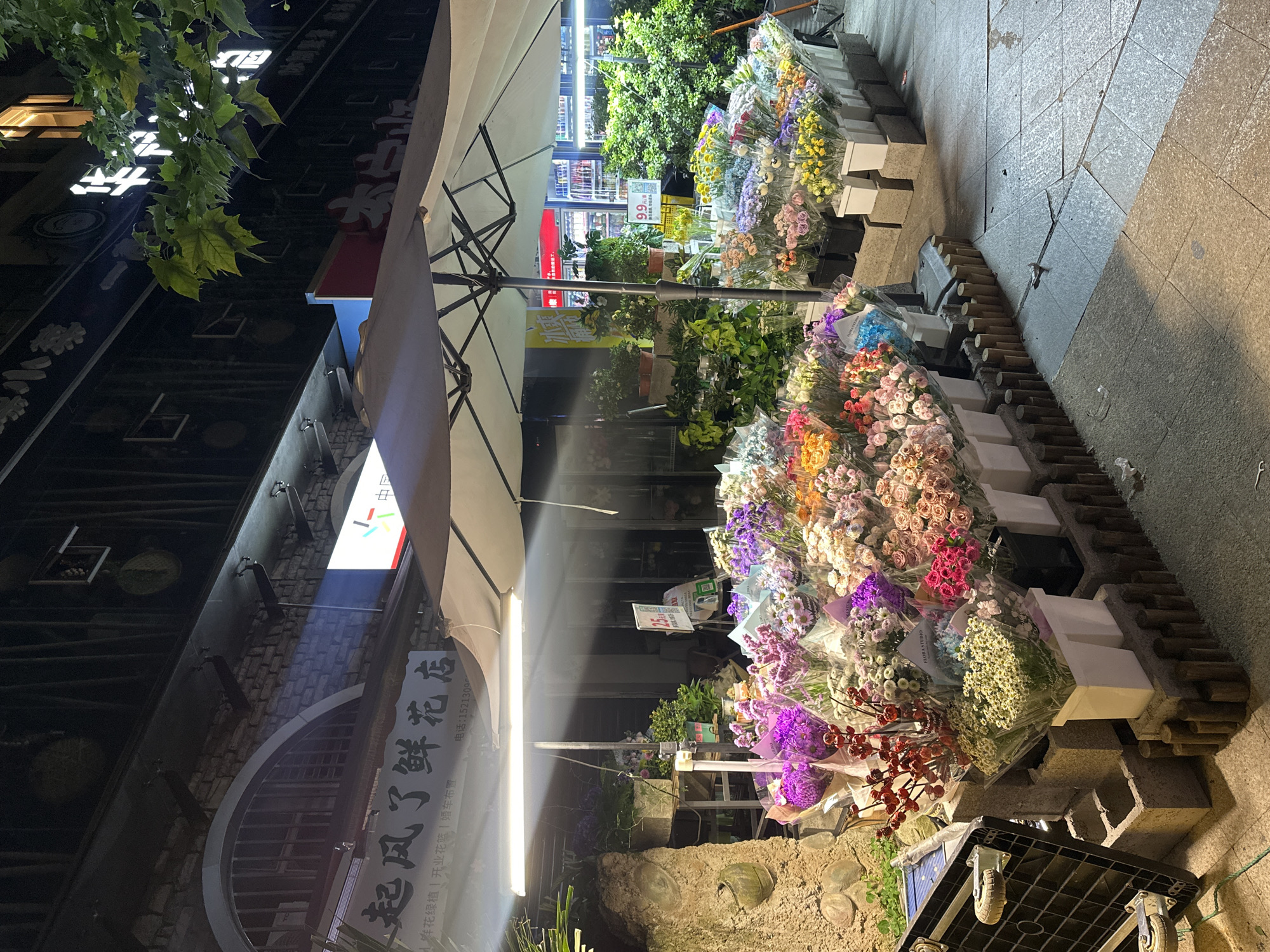 The image captures a street scene in Chongqing High-tech Zone, China, at night, featuring a flower shop. Numerous bouquets of various colors are displayed on tiered blocks, positioned along the edge of a pedestrian walkway made of square pavers. Above the flowers is a large, partially visible umbrella, providing shelter. Adjacent to the flowers is the flower shop's signage, reading "起风了鲜花店," along with details about their services, like floral arrangements and wedding car decorations. Further in the background is a brightly lit store, possibly a convenience store. Streetlights illuminate the area, and foliage from trees is visible in the upper left corner. A black rolling crate is located in the lower right corner.