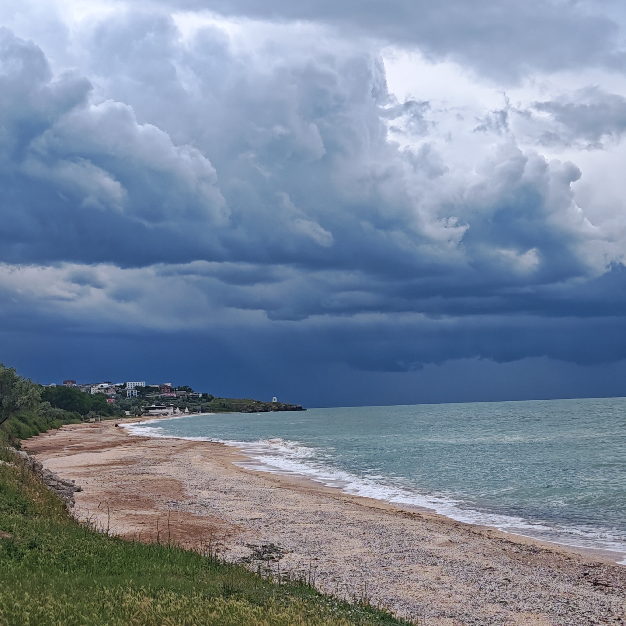 The image depicts a beach scene in городское поселение Щелкино, Ukraine, under a dramatic, stormy sky. Dark, heavy clouds dominate the upper two-thirds of the frame, casting a deep shadow over the scene.
Below the clouds, the water is a greenish-blue, with white-capped waves breaking gently on the shore. The beach itself appears to be a mix of sand and small stones.
Along the left edge of the frame, a grassy slope leads down to the beach. In the distance, along the shoreline, there are buildings and what appears to be a white gazebo or small structure on a point of land. The atmosphere suggests an impending storm or recent rainfall.