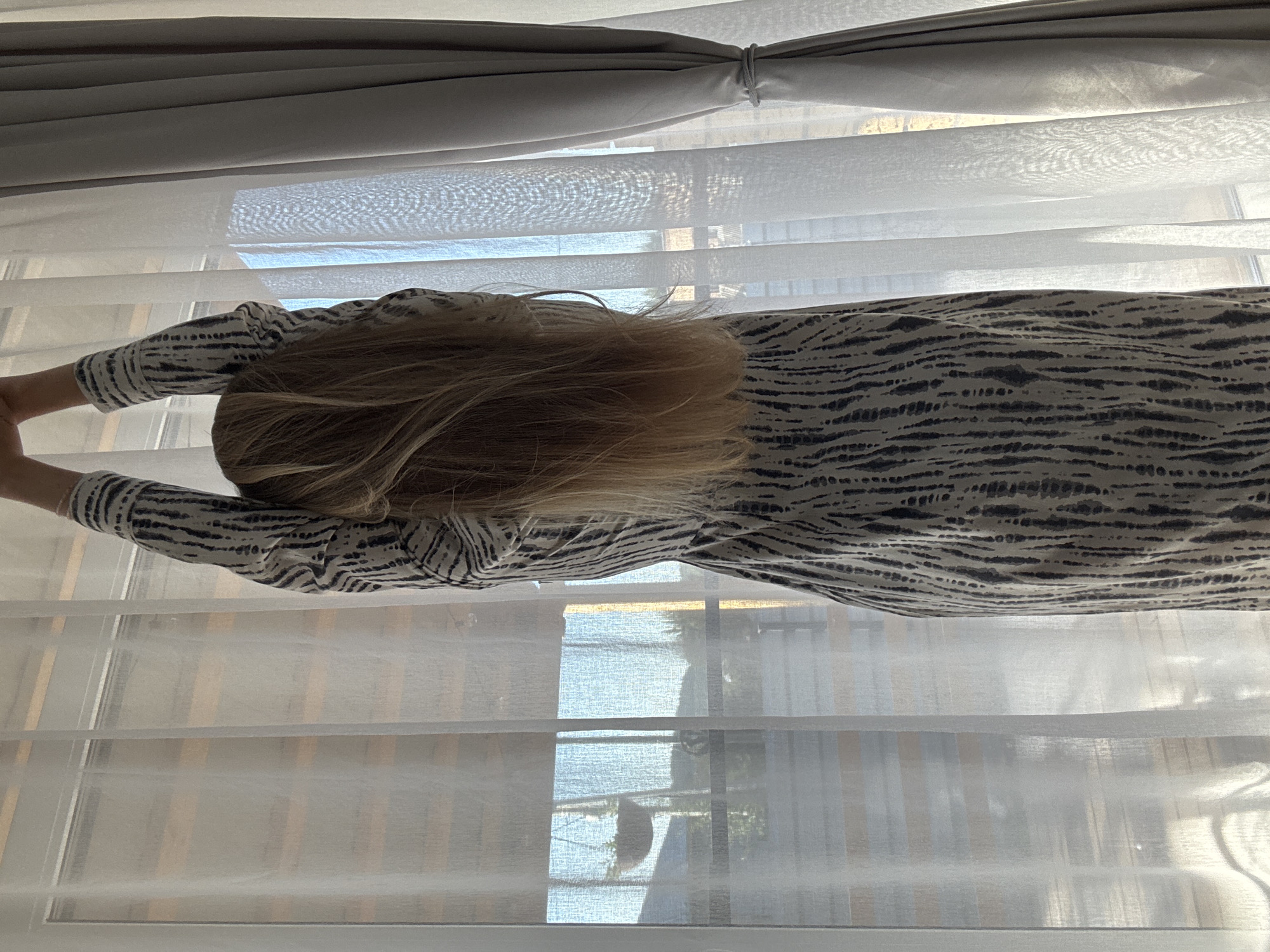 A person with blonde hair and wearing a black and white patterned long-sleeved shirt is standing in front of a window covered with white blinds and curtains. The location is Chaikovsky, Russia. The person's arms are raised, as if stretching or reaching for something. The blinds are partially open, allowing light to enter the room. Outside the window, blurred elements suggest a cityscape with buildings and trees. The image appears to be taken from a low angle.