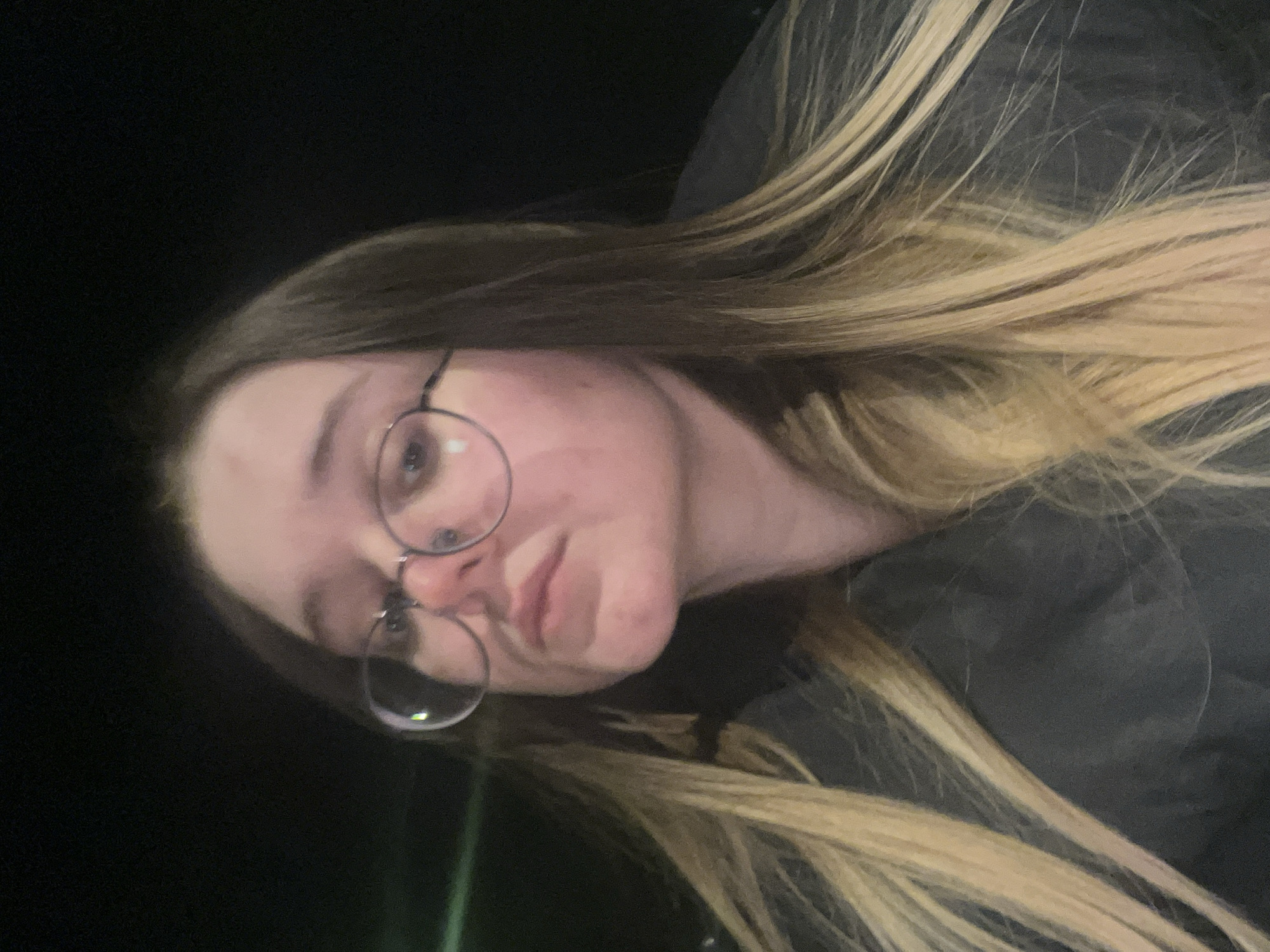 The image displays a close-up portrait of a young woman situated in the city of Pushchino, Russia. She possesses long, straight hair, which is dark brown at the roots and transitions to blonde highlights towards the ends. The woman wears round, wire-frame glasses. Her face is directed towards the camera with a neutral to slightly pouting expression, and her head is angled slightly upward. She is dressed in a dark, possibly black or dark grey, jacket or coat. The background is largely indistinct and dark, indicative of low light conditions, with a faint horizontal green light streak visible on the left side of the frame. No other individuals or significant objects are clearly discernible within the scene.