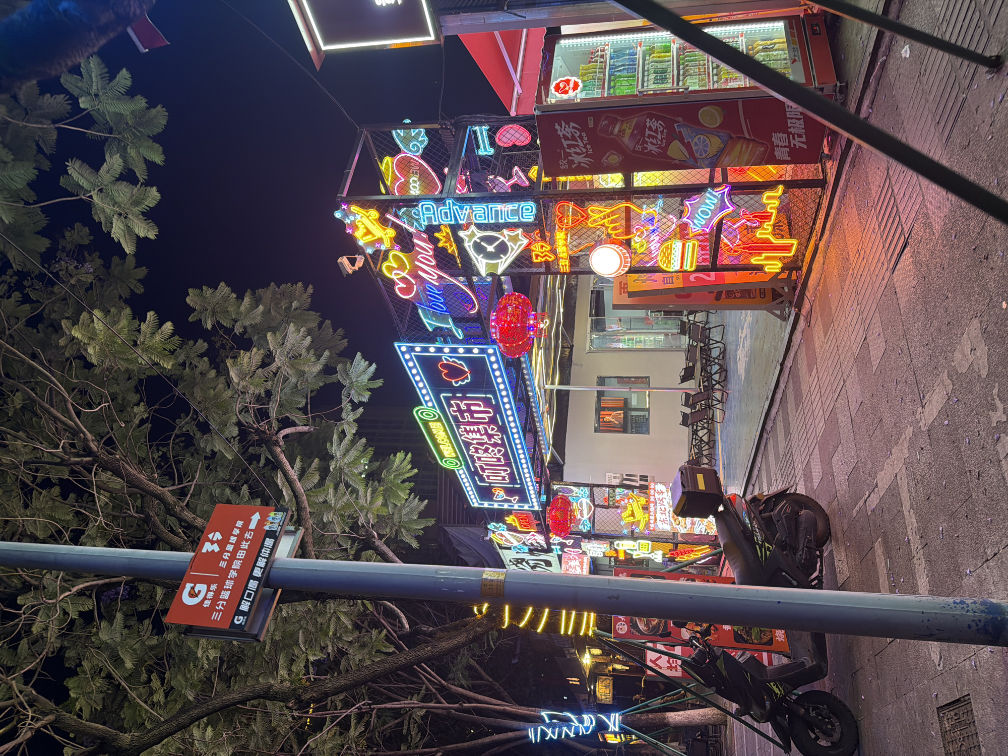 Here's a detailed description of the image:
**Location:** This photo was taken in the city of Chongqing High-tech Zone, China. This is determined by the signs and neon displays visible in the image.
**Environment:** The image captures a bustling urban scene at night, dominated by vibrant neon lights and signage. The setting appears to be a commercial area with shops and restaurants lining the street. The presence of trees and branches indicates an integration of greenery within the urban landscape.
**Objects:**
* A multitude of neon signs in various shapes, colors, and sizes advertise different businesses and products. Some of the signs include the words "Advance", "Welcome" in English, while others display Chinese characters.
* There is a shop or restaurant front visible in the background, with merchandise displayed behind a glass facade.
* A small electric scooter is parked on the side of the road.
* Streetlights and poles are also present, with signage attached to them in both English and Chinese.
* There is a small red sign showing directions.
* "MECO" and "Ice Tea" on drinks.
**Atmosphere:** The overall atmosphere is lively and energetic due to the abundance of lights and visual stimuli. The image captures the vibrancy and dynamic nature of urban life in Chongqing High-tech Zone.
