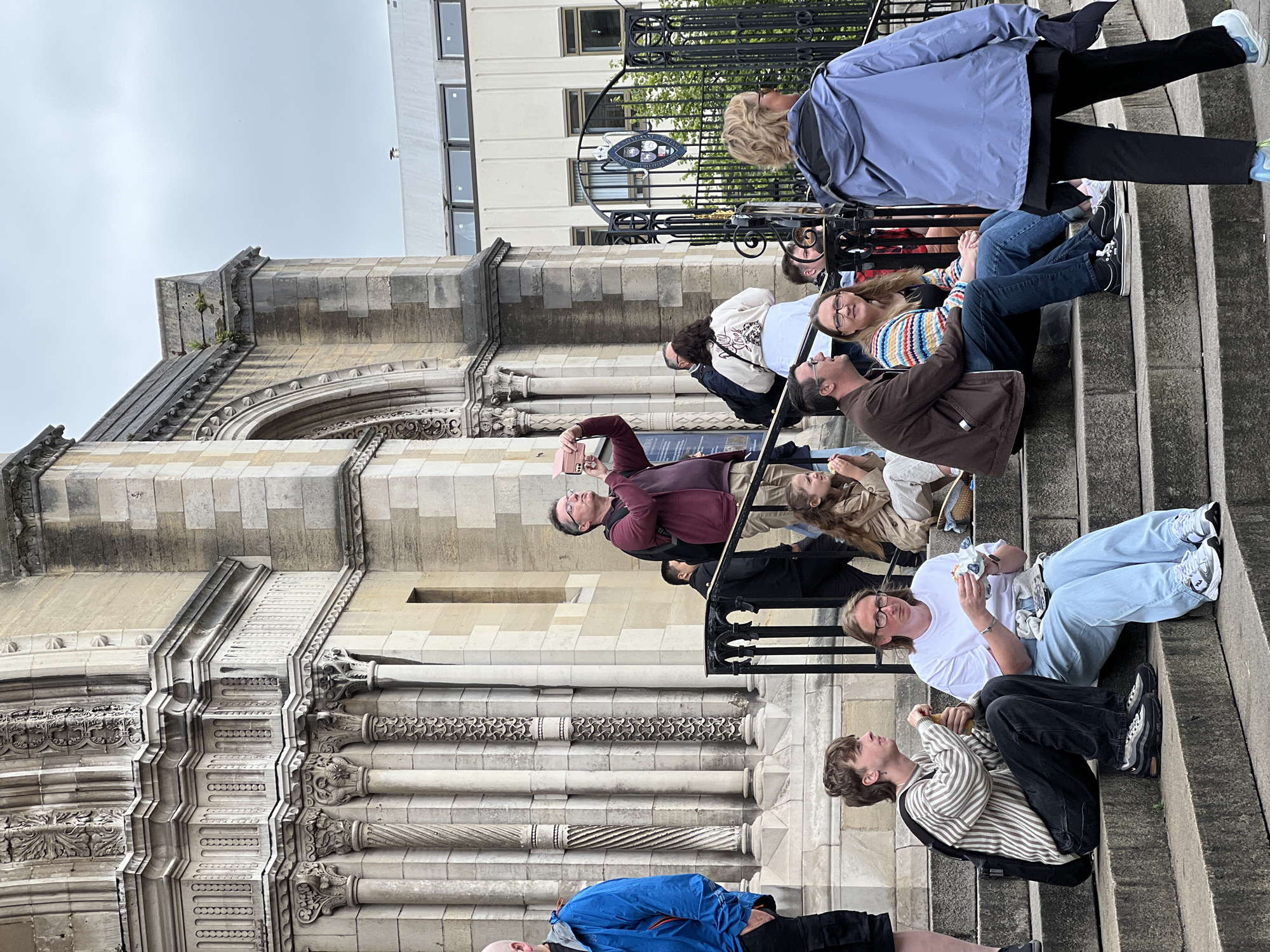 A group of people are gathered on the stone steps of a large, ornate building. The architecture suggests it is a historic or governmental structure. Several individuals are seated on the steps, some eating or looking at their phones. One man in the middle distance is holding up a phone, appearing to take a photograph. A woman in the foreground on the right is walking away from the camera, wearing a blue hooded jacket. The overall atmosphere appears casual, with people enjoying a day outdoors. The location is confirmed to be Belfast City District, United Kingdom.