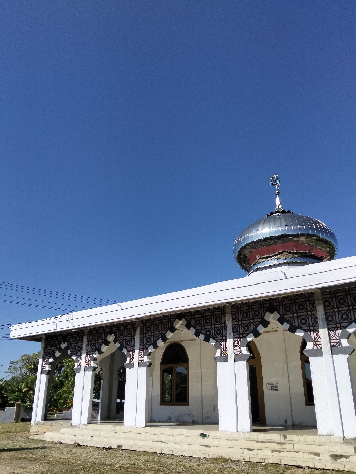 A mosque with a gleaming silver dome and a spire stands prominently against a cloudless blue sky. The building features a white facade with repeating arched entryways decorated with geometric patterns. A small rectangular window punctuates the wall. The mosque sits on a slightly elevated platform of stairs. The surrounding area appears to be a grassy field, suggesting a rural or suburban setting.