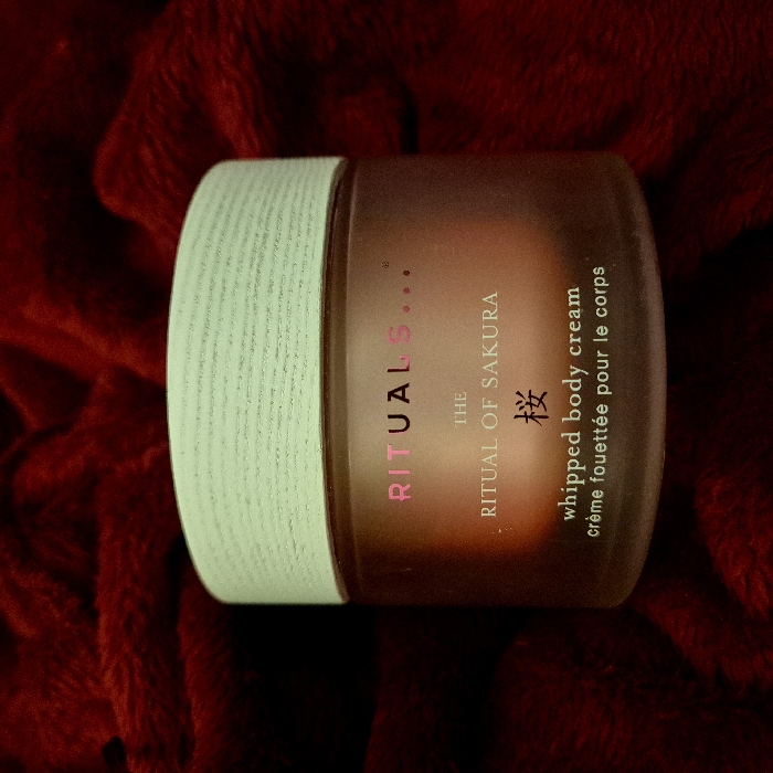 A jar of "Rituals..." whipped body cream, specifically "The Ritual of Sakura," rests on a textured, dark red fabric. The jar is brown with a light green lid featuring horizontal ribbed details. The words "Rituals..." are vertically aligned and in pinkish hue. Below that is written "The Ritual of Sakura" with an Asian character and "whipped body cream crème fouettée pour le corps" to the right.