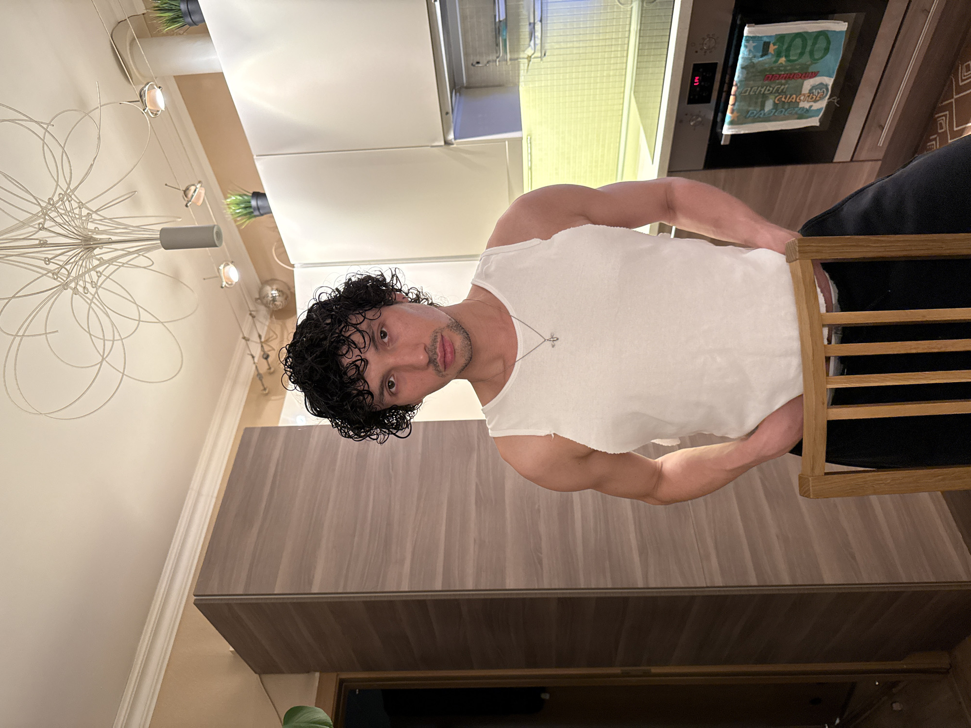 A man with curly dark hair, wearing a white tank top and a necklace, stands in a kitchen. He has a slight stubble and a neutral expression. Behind him, a modern kitchen is visible with white cabinets, wooden countertops, and a stainless steel oven. A patterned towel or cloth with "100" and Russian text is hanging from the oven door. A wooden chair with horizontal slats is partially visible on the right. The lighting suggests it is daytime or well-lit indoors.