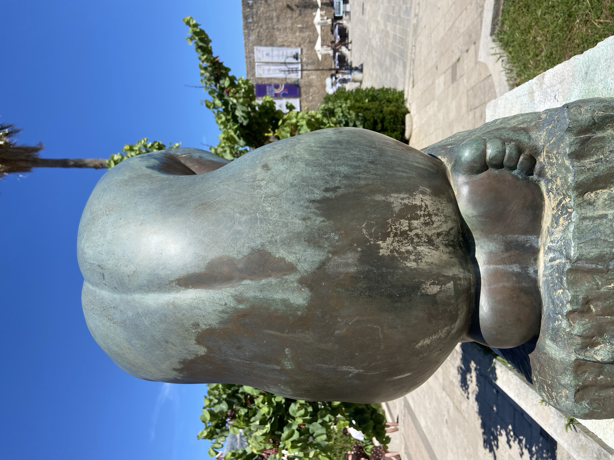 The image captures a bronze sculpture in Budva, Montenegro. The sculpture features an abstract human form with a prominent rounded belly or torso resting on a stylized foot. The metal surface exhibits a patina with variations in color and texture, suggesting age and exposure to the elements. In the background, a stone building is visible, with a couple of banners on the wall. Tables and chairs suggest a cafe or outdoor seating area. The sky is bright blue, indicating a sunny day. The setting appears to be a public square or pedestrian area in the city.