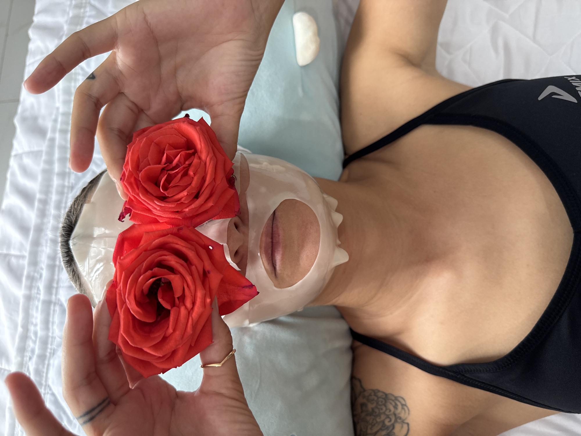 A close-up aerial shot shows a person lying down with a face mask on. Two red roses are held in front of the person's eyes, obscuring them. The person is wearing a black sports bra. A light blue pillow is visible behind the person's head. The background is a white quilted surface, possibly a bed. There are tattoos visible on the person's left arm and hand. The scene appears to be indoors, in a domestic setting, likely a bedroom. There is no information to suggest this image is located in Nha Trang, Vietnam.