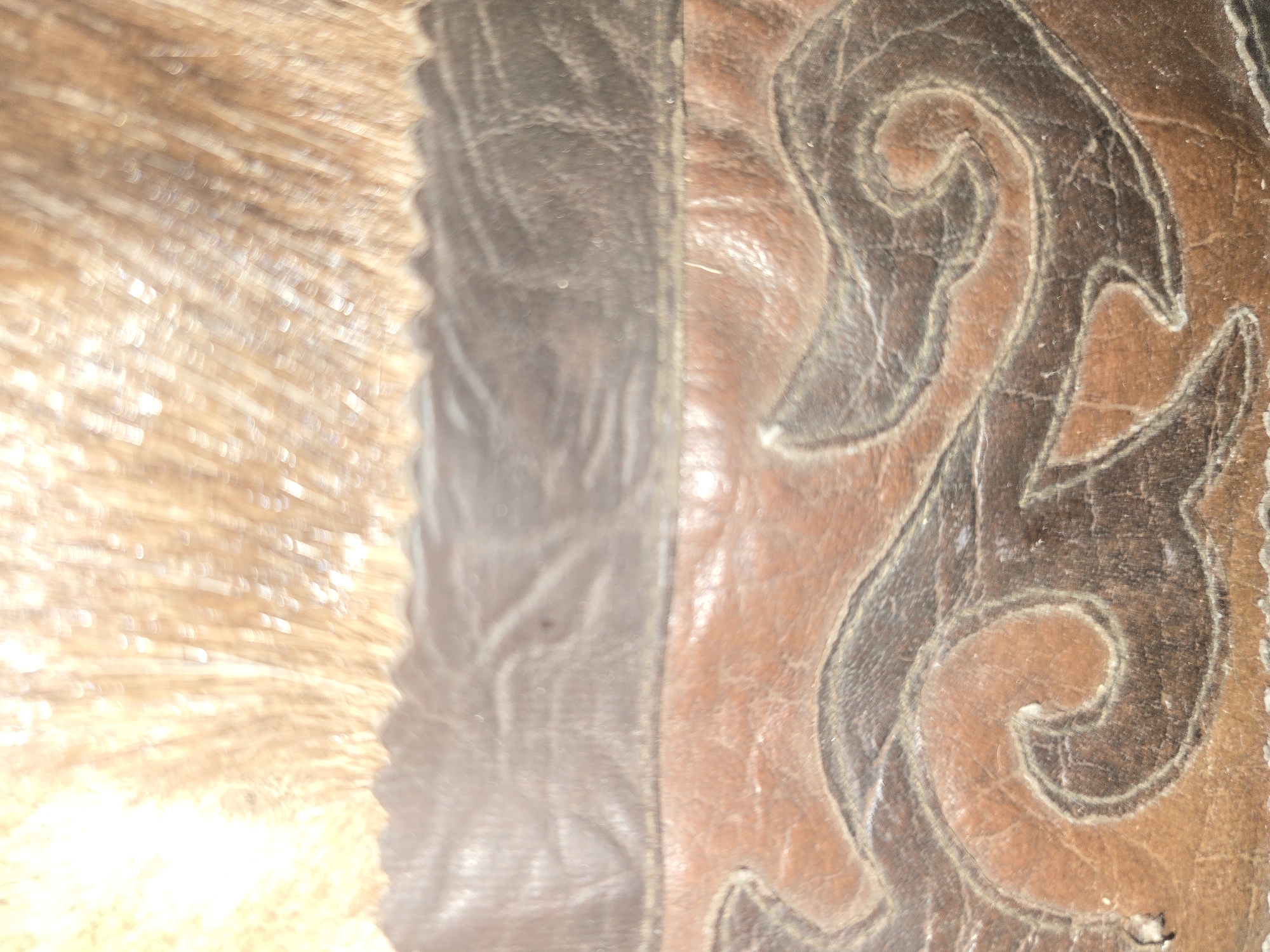 The image shows a close-up of a decorative leather item, possibly a bag or a piece of furniture. The top portion of the image features a fuzzy, light-colored material that appears to be fur or thick yarn, with a saw-tooth edge along the bottom. Below this, there is a section of dark brown leather with a textured, slightly wrinkled surface. The bottom section of the image showcases a patterned design on lighter brown and dark brown leather. The pattern consists of swirling, abstract shapes that resemble waves or stylized foliage, with contrasting dark lines accentuating the curves. The overall impression is one of rustic craftsmanship and natural materials.
Without any contextual information or recognizable landmarks, it is impossible to determine the location within the city of Ulan-Ude, Russia, or any other specific geographical setting.