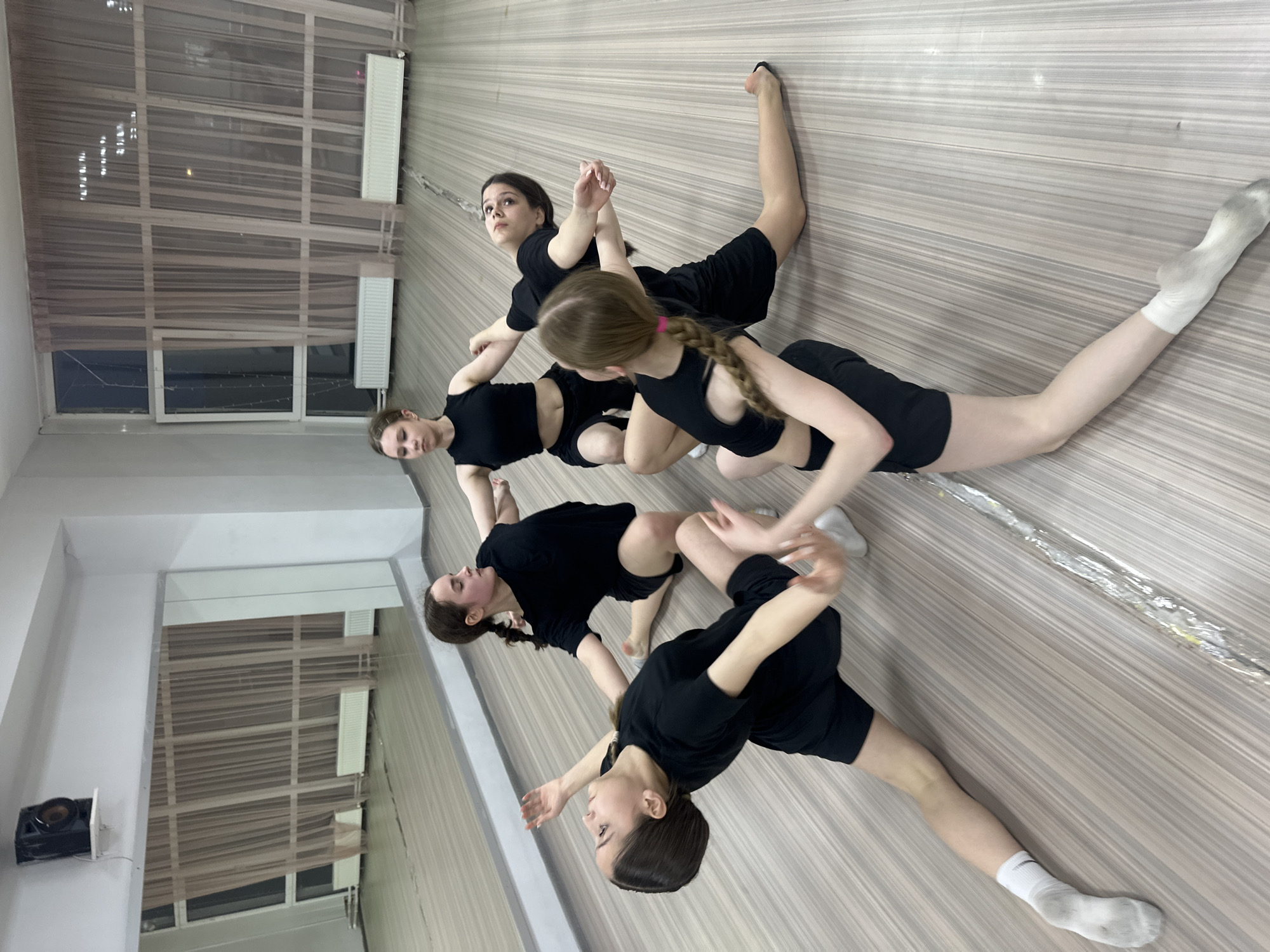 This image captures five young women, likely in their late teens or early twenties, engaged in a synchronized dance, stretching, or exercise routine on a light wooden floor. All are dressed uniformly in black athletic short-sleeved tops and shorts, and some wear white socks. They are positioned on the floor in various flexible poses, demonstrating concentration and control, with their expressions appearing focused and calm. One woman in the foreground has a long braid, while others have their hair pulled back or styled differently.
The setting is an indoor studio, possibly a dance or fitness studio in Votkinsk, Russia. The room features a clean, light-colored wooden floor with distinct horizontal planks. Large windows, covered by sheer, light-colored curtains, dominate one wall, beneath which two white radiators are mounted. A projector is visible mounted high on a wall in the background, and a large reflective surface, likely a mirror, runs along another wall, reflecting parts of the room. A long, cylindrical object, wrapped in clear plastic, lies on the floor. The image is taken from an elevated, slightly angled perspective, looking down at the participants. No text is visible within the frame. The lighting suggests daytime, though internal artificial light is also present.