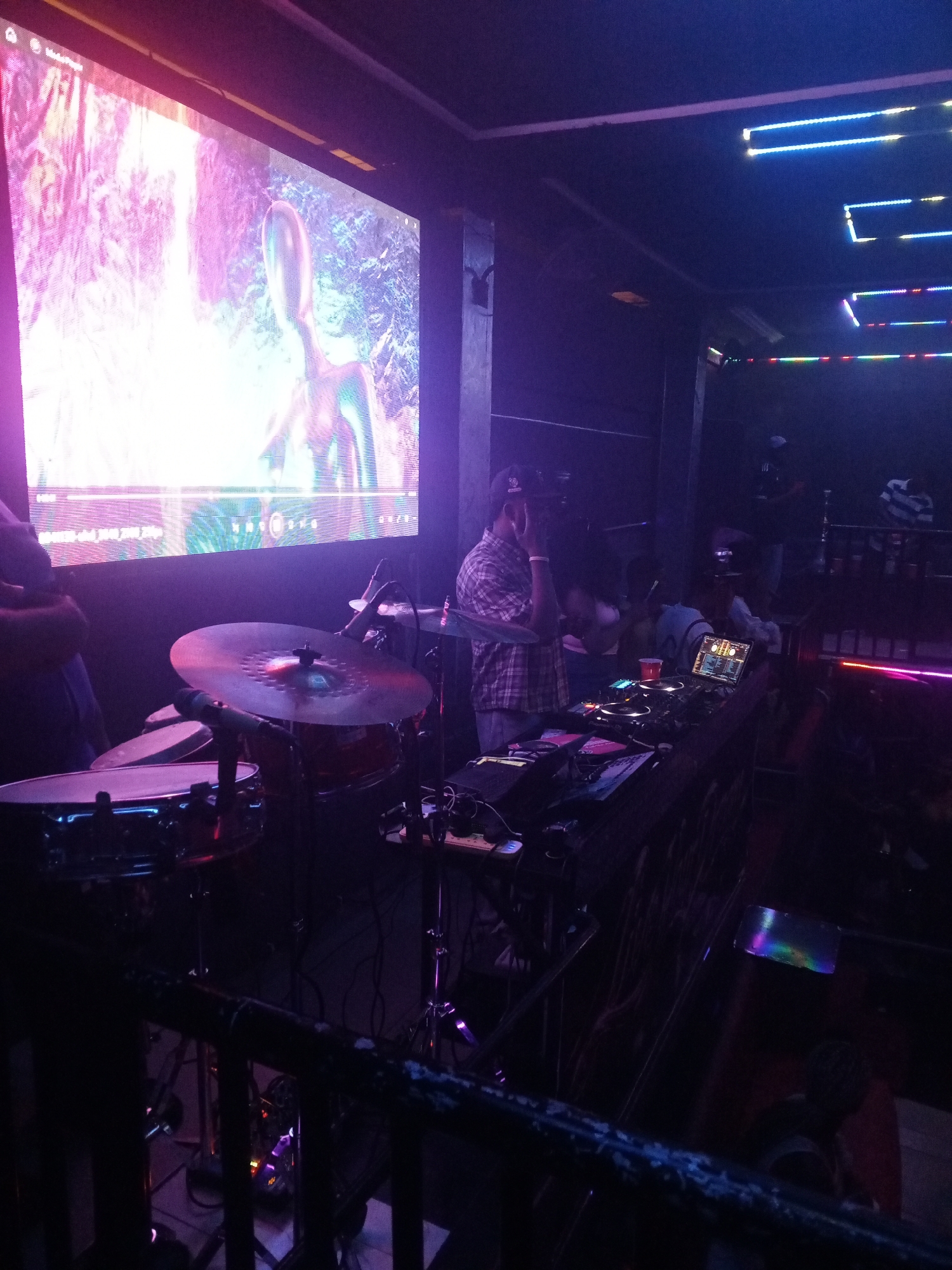 A person is operating a DJ setup in a dimly lit environment. A drum set is present on the left. A large screen displays a video. Other people are in the background near a bar. The location is a club in an unknown city.