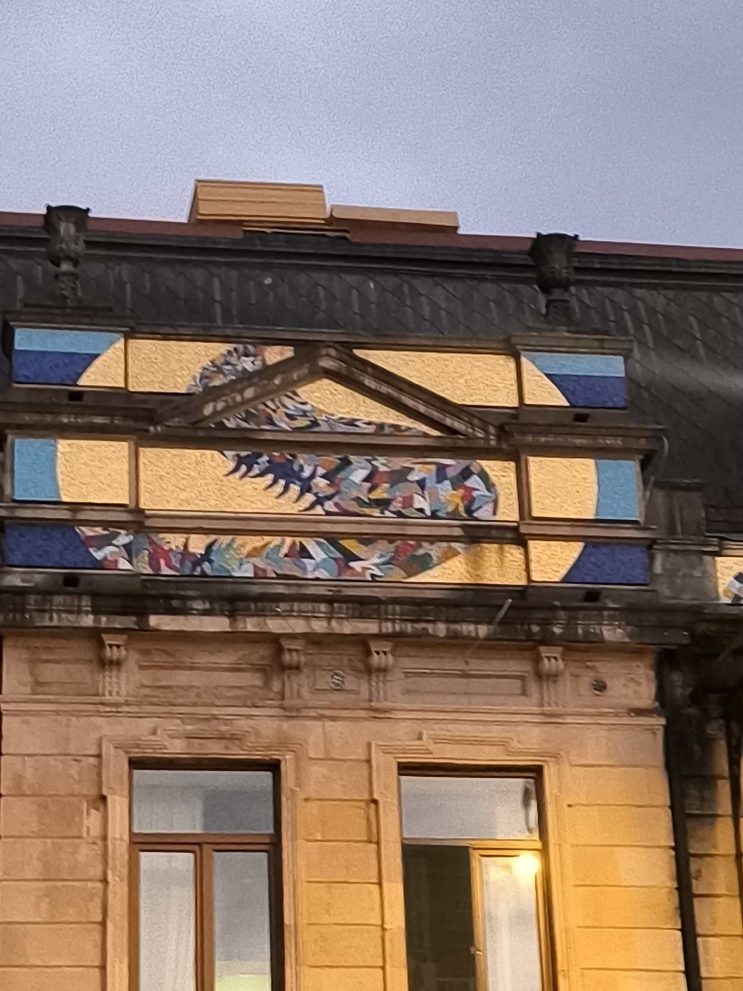 The image shows an exterior architectural detail of a building in Batumi, Georgia. The upper portion of the facade features a decorative mosaic panel. This mosaic is set within a golden, rectangular frame and depicts an abstract, feather-like motif in various colors including blue, white, red, and green. The mosaic is flanked by rectangular sections of blue and golden tiles, with curved segments of blue tiles on the far left and right, resembling stylized suns or moons. Above the mosaic, the dark, sloped roof of the building is visible, with some wooden planks resting on its edge. Below the mosaic panel, a stone facade with decorative moldings and two windows with wooden frames can be seen. The windows are illuminated from within, casting a warm glow. The sky above is overcast and grey.