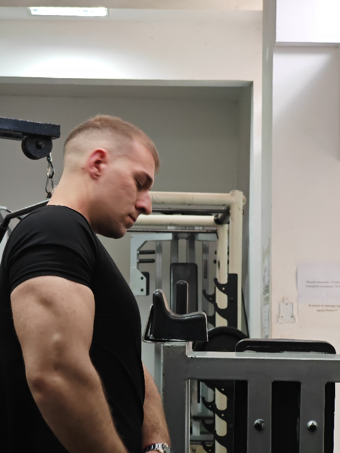 An indoor, eye-level photograph captures an adult male within a gym facility located in Shumen, Bulgaria. The subject is positioned in profile, facing right, with his head inclined downwards and eyes partially closed, suggesting a moment of rest or focus. He has a muscular build, short light-colored hair styled in a fade, and wears a black short-sleeved t-shirt. A silver-colored metal watch is visible on his left wrist. The man stands adjacent to a grey metal gym apparatus featuring a black padded rest in the immediate foreground. In the background, other gym equipment is visible, including a cable machine with a black pulley and chain in the upper left quadrant, and several weight stack machines with metal frames. The facility's walls and ceiling are light-colored. A linear fluorescent light fixture is present on the ceiling. On the right-hand wall, a white rectangular sign with black Cyrillic text is affixed, providing information such as "Месечен абонамент: 22 лв." (Monthly subscription: 22 BGN), "Еднократно посещение: 3 лв." (Single visit: 3 BGN), and a rule regarding appropriate attire: "В залата за тренировки само в чисто и сухо спортно облекло!!" (In the training hall only with clean and dry sports clothing!!), confirming the location as a fitness center in Bulgaria.
