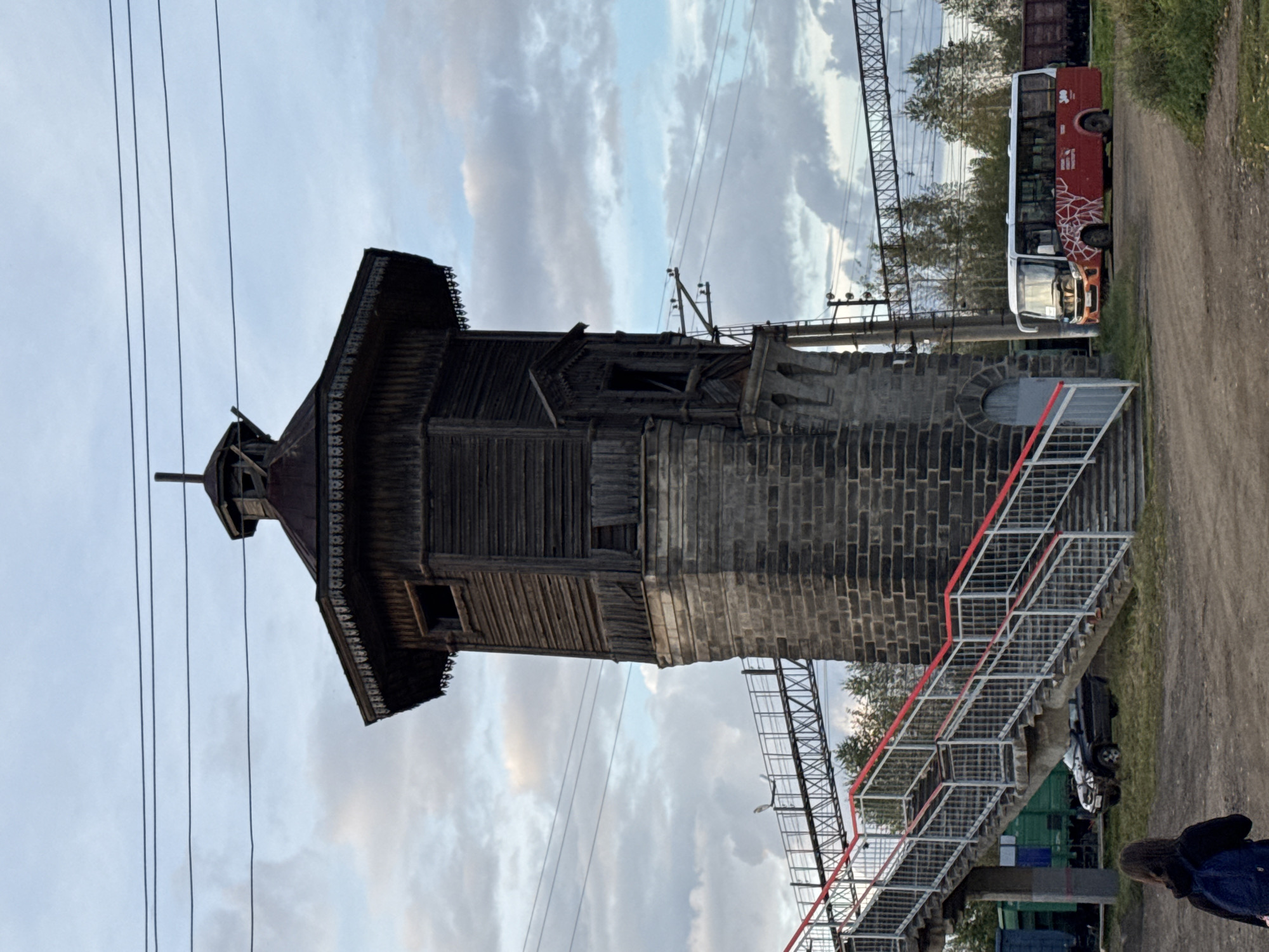 This is a full, eye-level, outdoor shot of an old wooden water tower at a train station in Komarikha, Russia. The sky is cloudy with patches of blue. The tower is constructed of rough-hewn dark wood planks on a stone base, and it has a multi-tiered roof. In front of the tower is a pedestrian overpass with a red railing and metal stairs leading up. A red and white bus is parked to the right of the tower, facing left. A black SUV is parked further back, to the left of the overpass. A young person wearing a backpack is standing in the foreground, facing away from the camera. Train tracks and freight cars are visible in the background, along with trees.