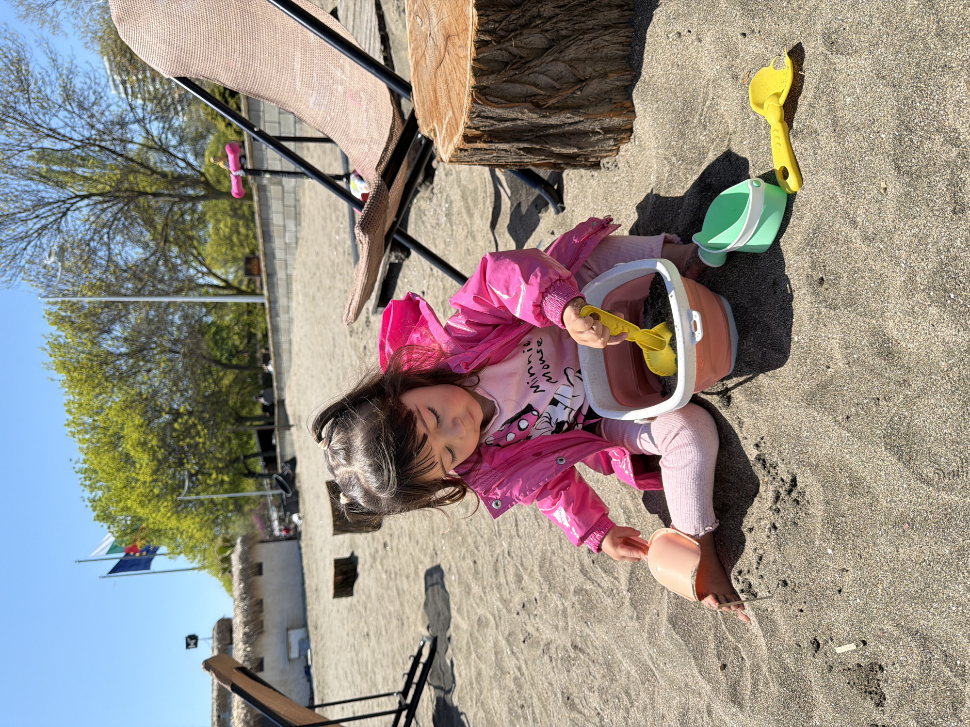 A young child, possibly a girl, is sitting on a sandy beach next to beach chairs and a tree stump. She is wearing a pink jacket over a pink shirt with a cartoon graphic, pink leggings, and is playing with beach toys, including a bucket, a green pail, and a yellow shovel. The child is interacting with the bucket, holding a yellow shovel. The environment includes sand, beach furniture, and a tree stump. A stone wall, flags, and trees can be seen in the background under a clear sky. The location is a beach in the city of Unknown, Unknown.