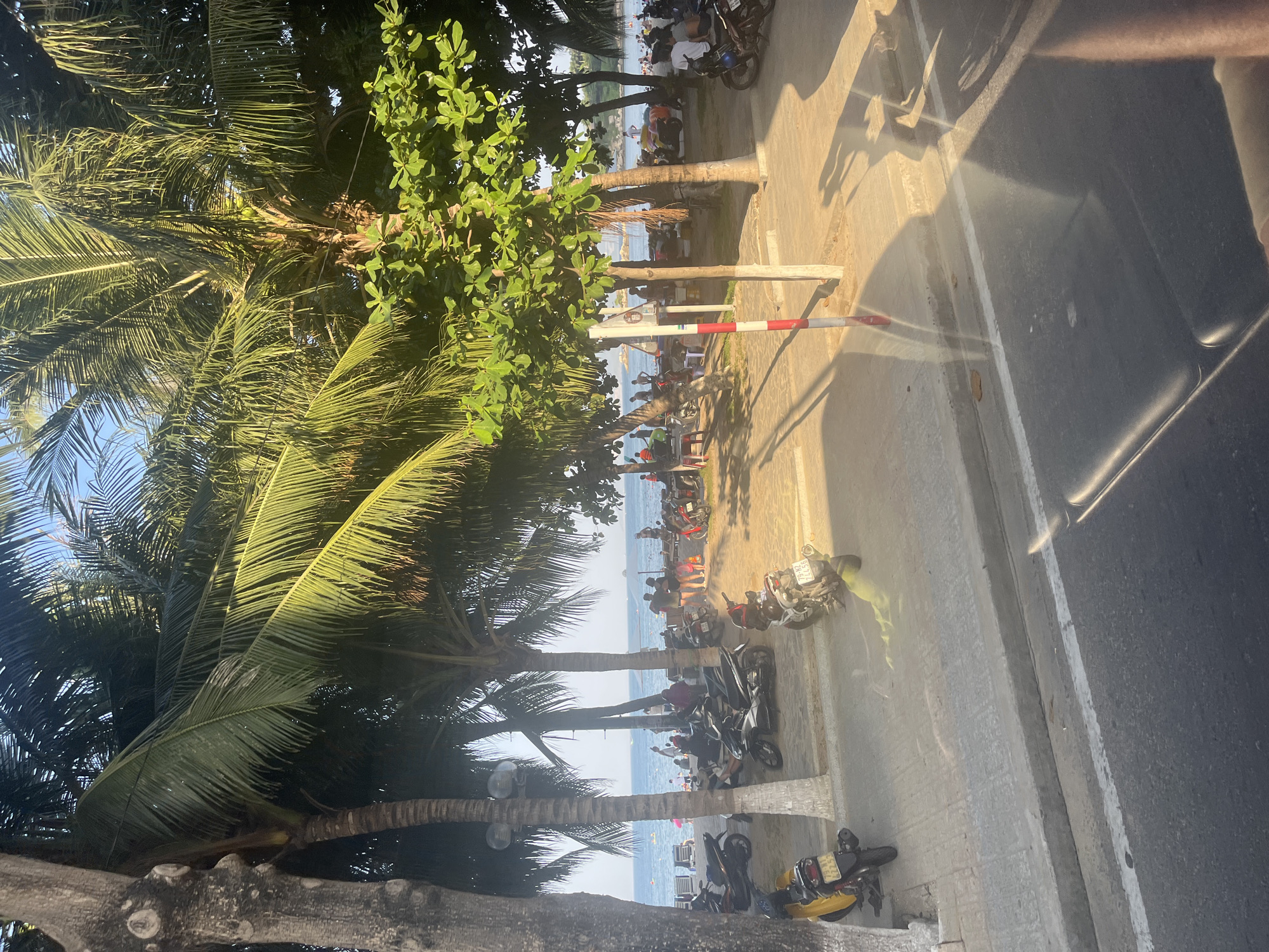This is a photograph taken on a sunny day in Nha Trang, Vietnam, likely from a moving vehicle, looking towards a beach and the sea. In the foreground, a grey asphalt road with a white line is visible, and a concrete curb separates it from a sidewalk. The sidewalk is paved with grey concrete and has a textured surface. Several parked motorcycles, predominantly scooters, are visible on the sidewalk and the adjacent sandy area. One yellow scooter is parked on the left, and a white and black scooter is further down. In the middle ground, a beach lined with palm trees separates the sidewalk from the ocean. People are scattered on the beach, some standing, some sitting on beach chairs, and others walking. The sea is a bright blue with gentle waves, and there are a few small boats in the distance. The sky is clear and blue. The presence of palm trees, beachgoers, and the ocean suggests a coastal resort town, consistent with Nha Trang. The overall scene depicts a bustling beachside area with common modes of transportation.