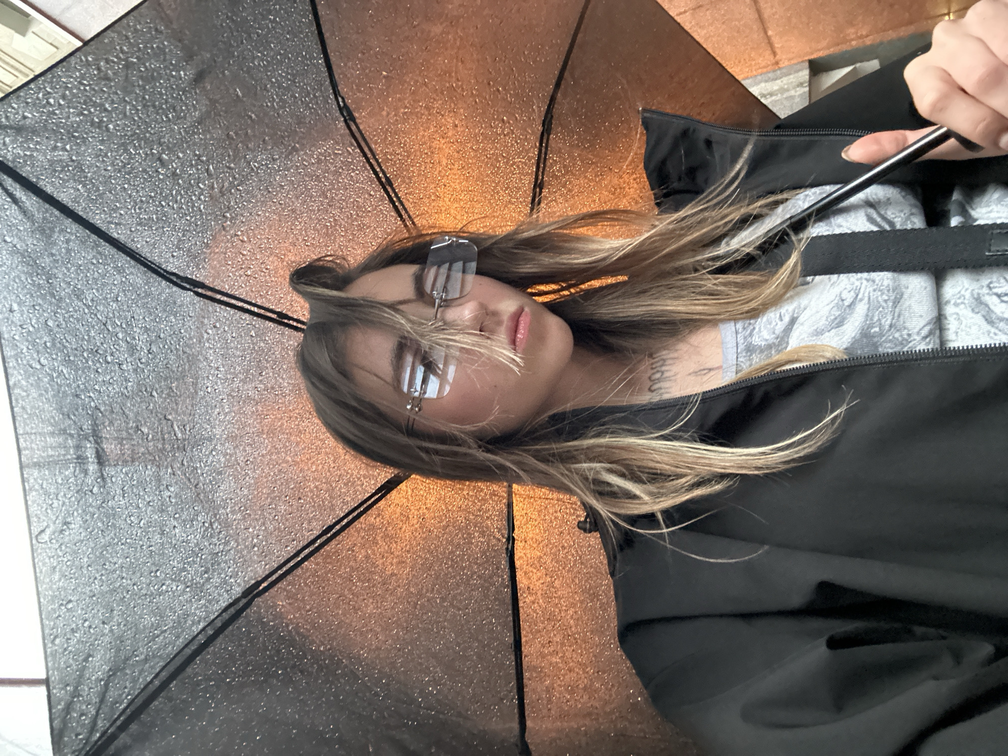 A young woman is captured in a selfie, holding a black umbrella with water droplets on it. She is wearing a black jacket over a grey t-shirt. Her hair is long and blonde, partially covering her face and glasses. She is looking down with a neutral expression. The background is blurred, with an orange glow suggesting artificial lighting. The image appears to be taken outdoors in what could be a city environment. There is no clear indication of her location in Moscow, Russia, or any specific details about the environment that would confirm it.