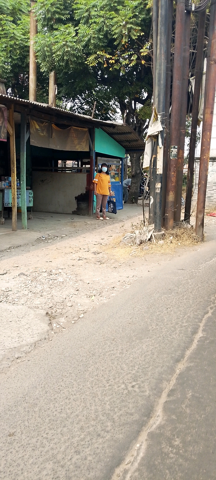 A person wearing an orange shirt and brown pants stands on a dirt road in front of a small roadside stall. The stall has a corrugated metal roof and a blue facade. The person is wearing a blue face mask. In the background, there are trees and utility poles. The road is unpaved and dusty. The scene appears to be in an urban or semi-urban area. The image is tagged with "Bekasi, Indonesia," suggesting the location.